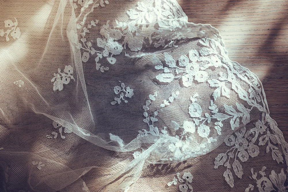 Close-up of delicate white lace wedding dress fabric with floral embroidery, partially draped on a wooden surface.