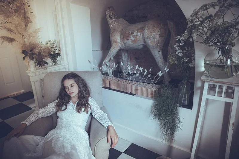 A bride with long brown hair in a white wedding dress relaxing in a beige armchair in a room decorated with plants, flowers, and a large abstract horse sculpture.