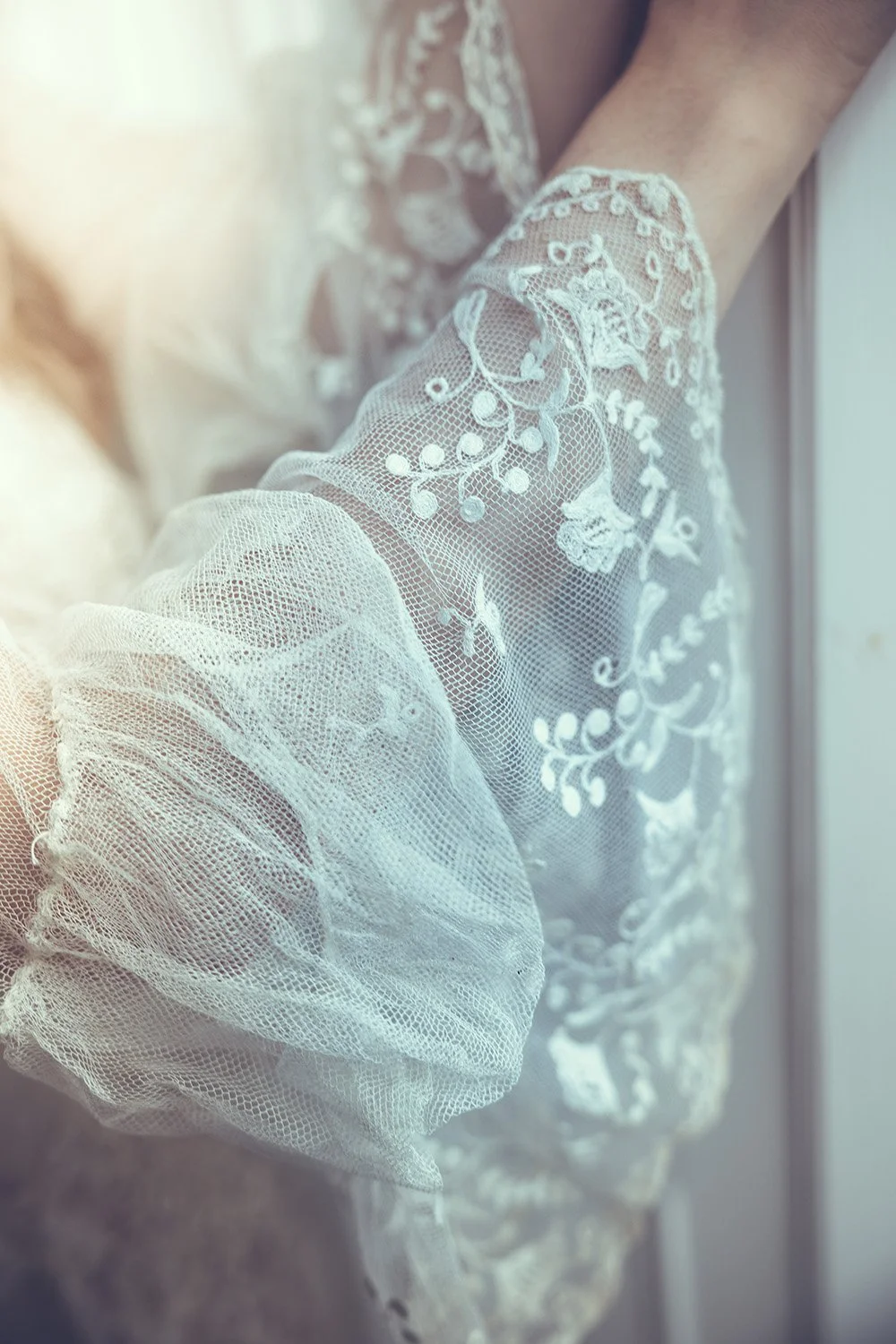 Close-up of sheer, embroidered lace fabric with floral design, in off-white color, draped with light illuminating it.