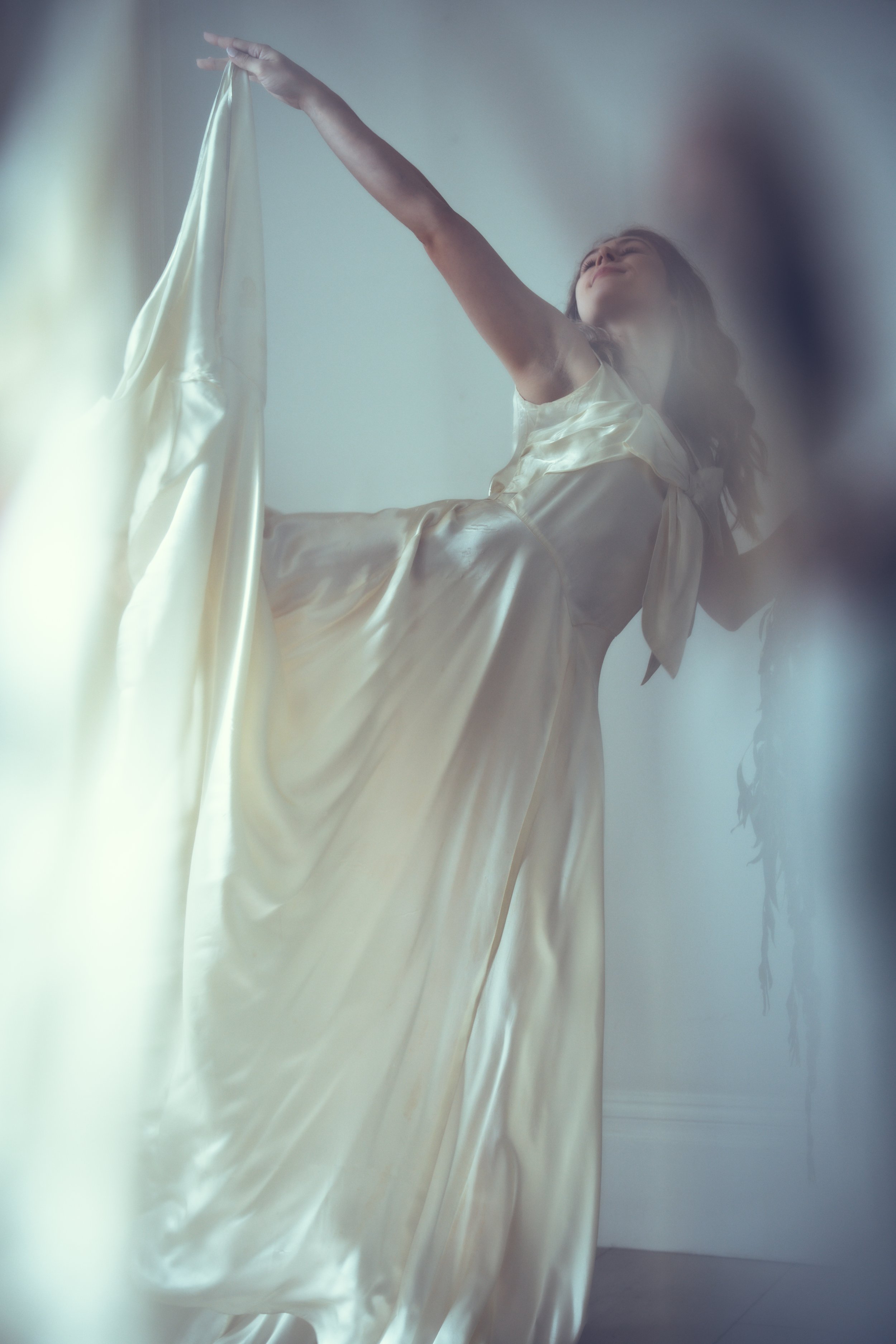 A bride in a flowing, silky, cream-coloured wedding dress reaching upward, with a soft, blurred background.