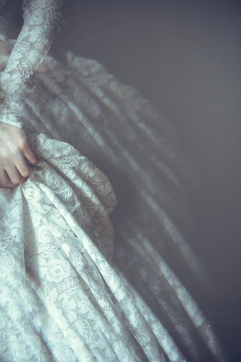 Close-up of a person wearing a white lace dress, with their hands gently clasped together, against a dark background.