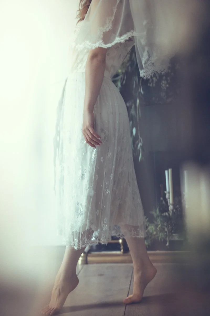 A bride-to-be in a white lace Tuesday's Bride wedding dress standing near a mirror, with her hand resting on her hip, while her reflection can be seen on the mirror.