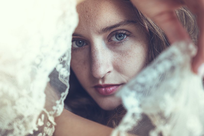 A bride with blue eyes and freckles gazes through lace wedding fabric.