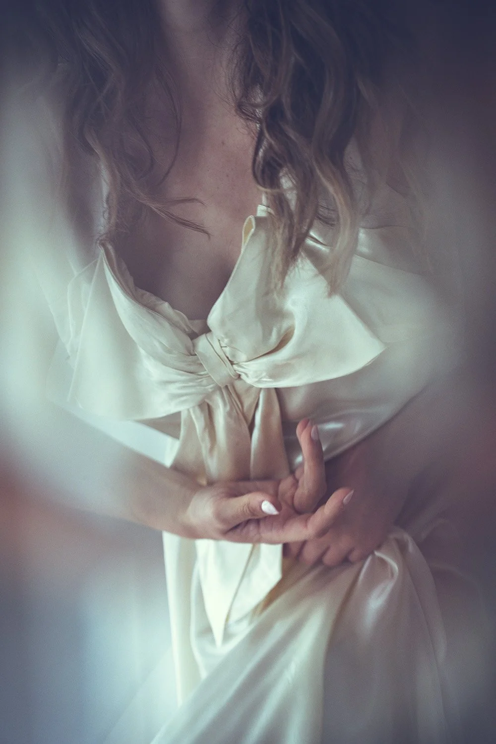 A bride with wavy hair wearing a satin cream-coloured Tuesday's Bride wedding dress with a bow, seen through a blurry or foggy lens.