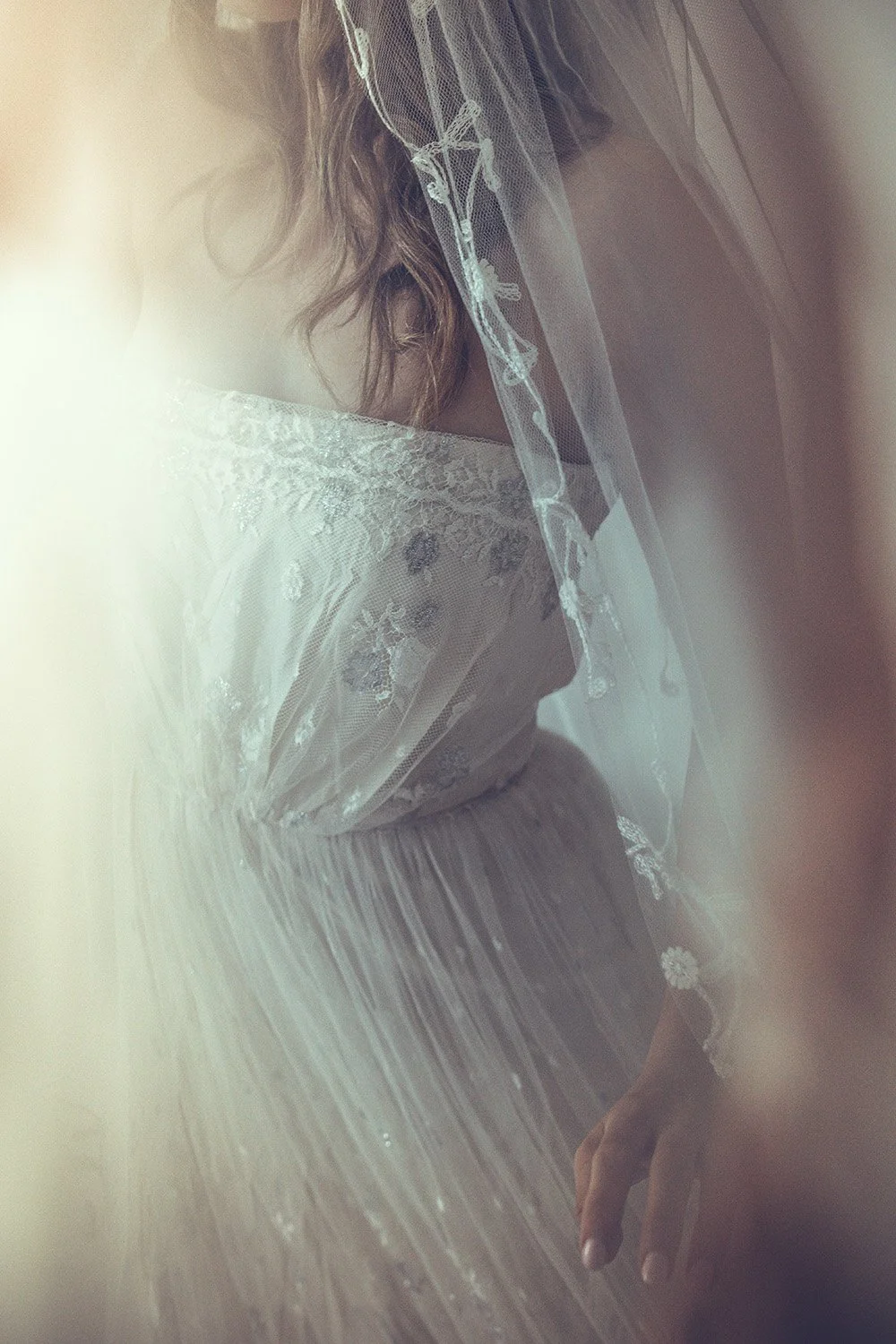Close-up of a woman in a delicate white lace wedding dress with a sheer veil, captured in soft, diffused light.