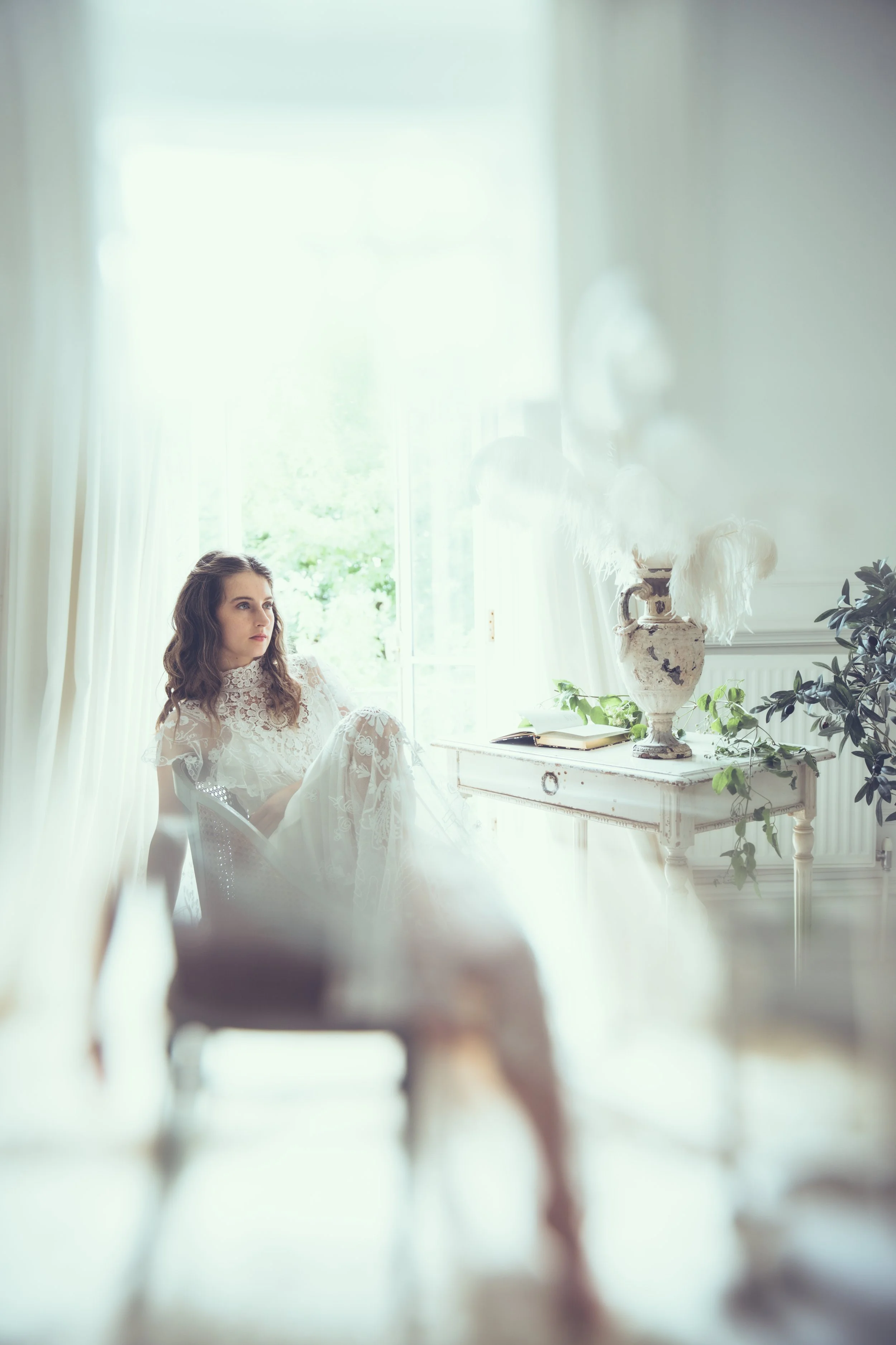 A bride in a white lace Tuesday's Bride wedding dress relaxing indoors near a window, with a table holding a vase, book, and plants, in a bright, airy room.