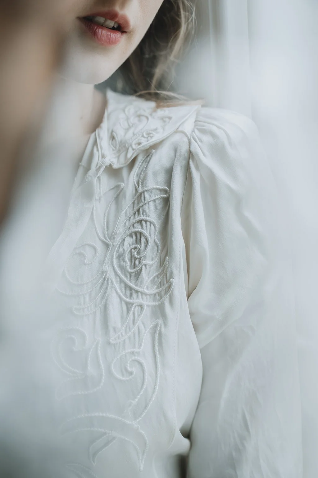 Close-up of a woman's face and shoulder, wearing a white embroidered wedding dress with intricate floral patterns, soft natural light.