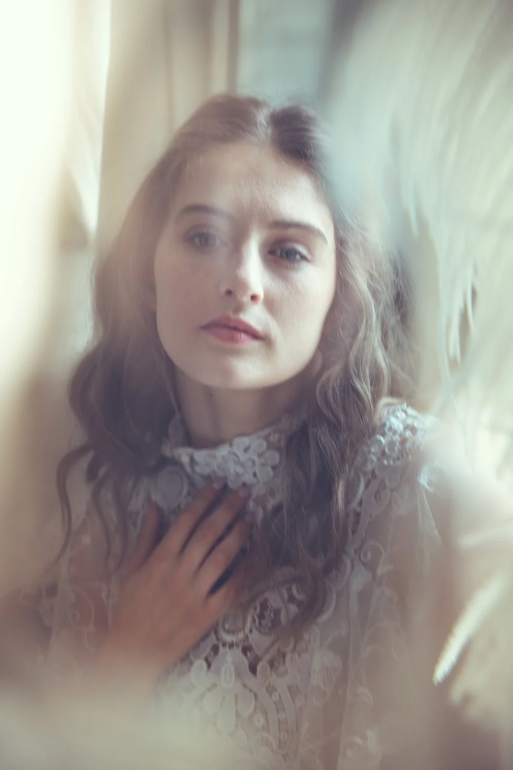 Abride-to-be with wavy brown hair and blue eyes, wearing Tuesday's Bride wedding dress, looking out of a window with soft natural light and a contemplative expression.