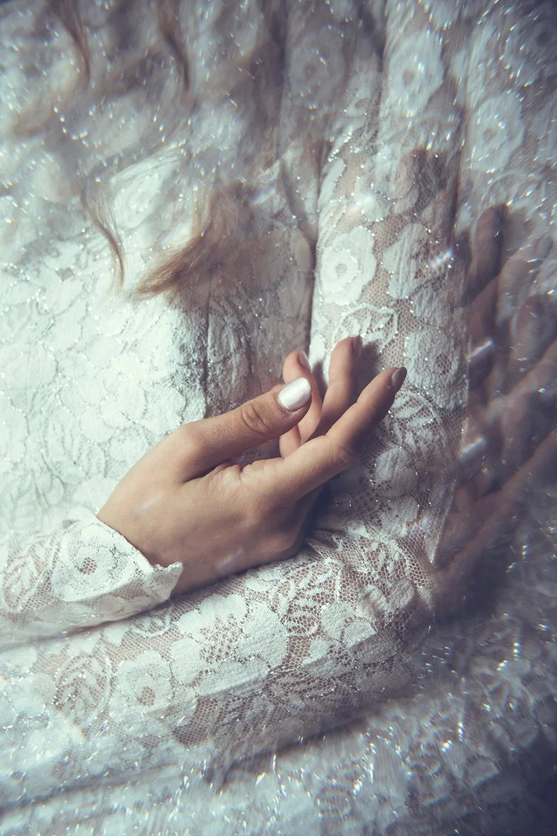 A close-up of a bride's hand with a white painted nail, touching their lace wedding dress sleeve with reflections and ripples distorting the scene.