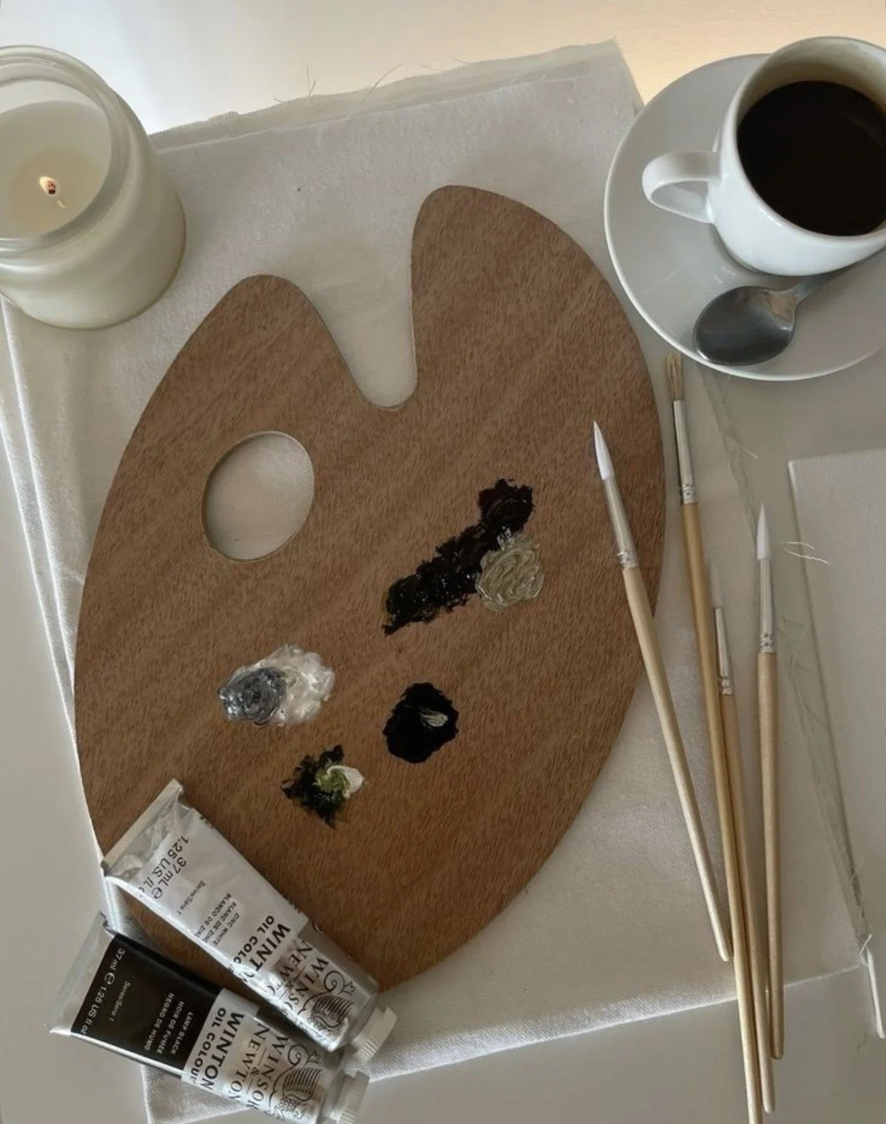 An artist's palette with various dollops of paint, a paintbrush, and paint tubes on a table next to a cup of coffee, a lit candle, and balancing spoons. A piece of Cintia Fauszt Design Studio.