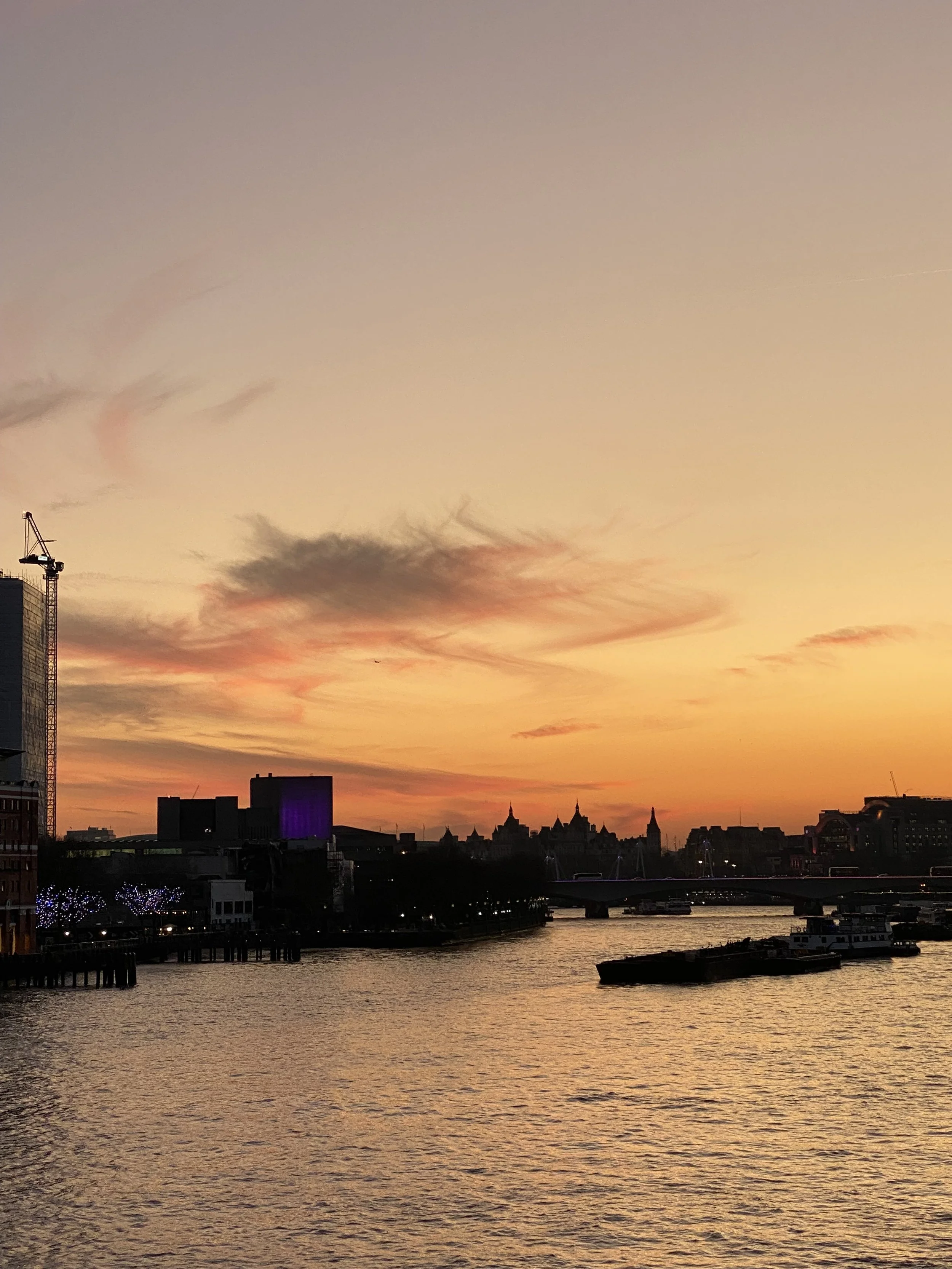Sunset over a city skyline with water in the foreground, including boats and illuminated buildings in London.