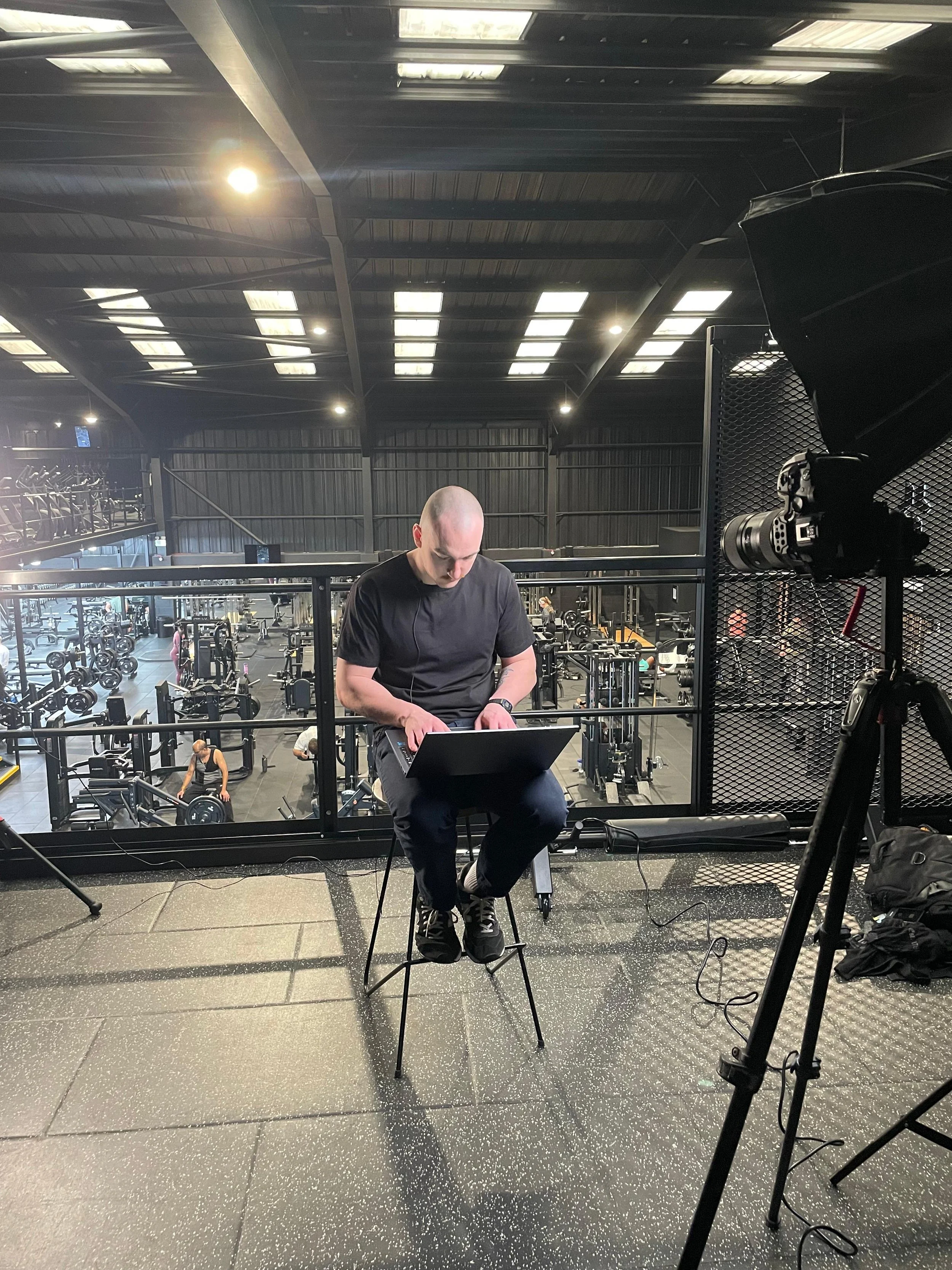 A man in a black shirt sitting on a stool with a laptop, recording a video with a camera on a tripod inside a gym.