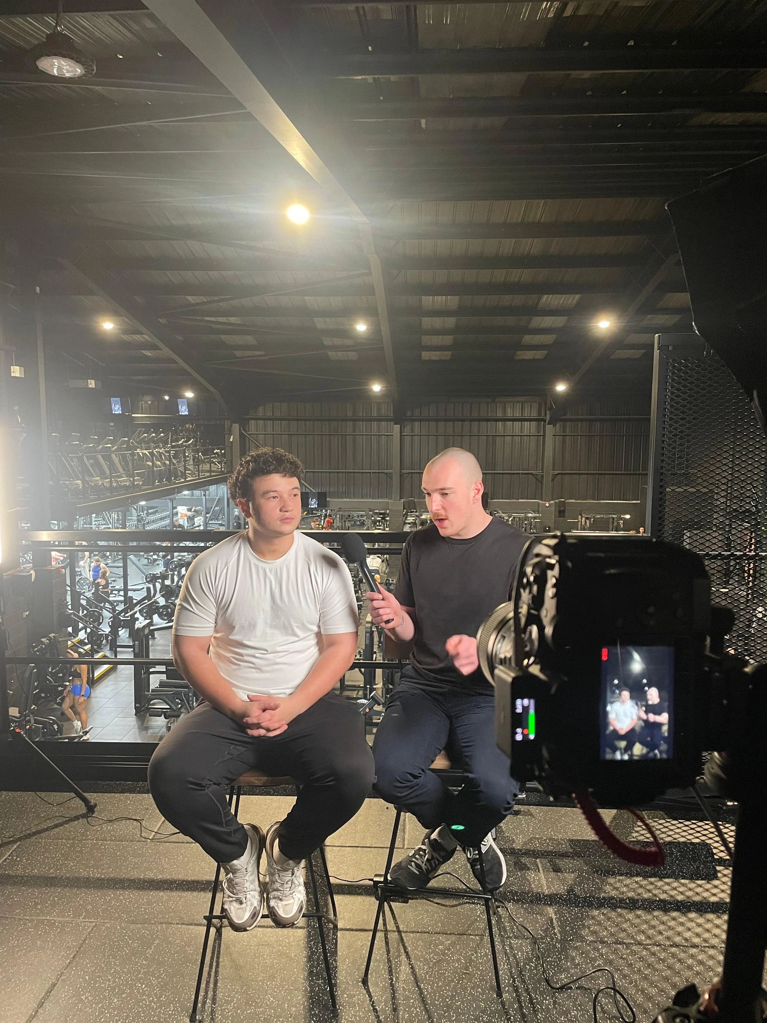 A man is recording an interview with a young man in a gym with fitness equipment in the background.