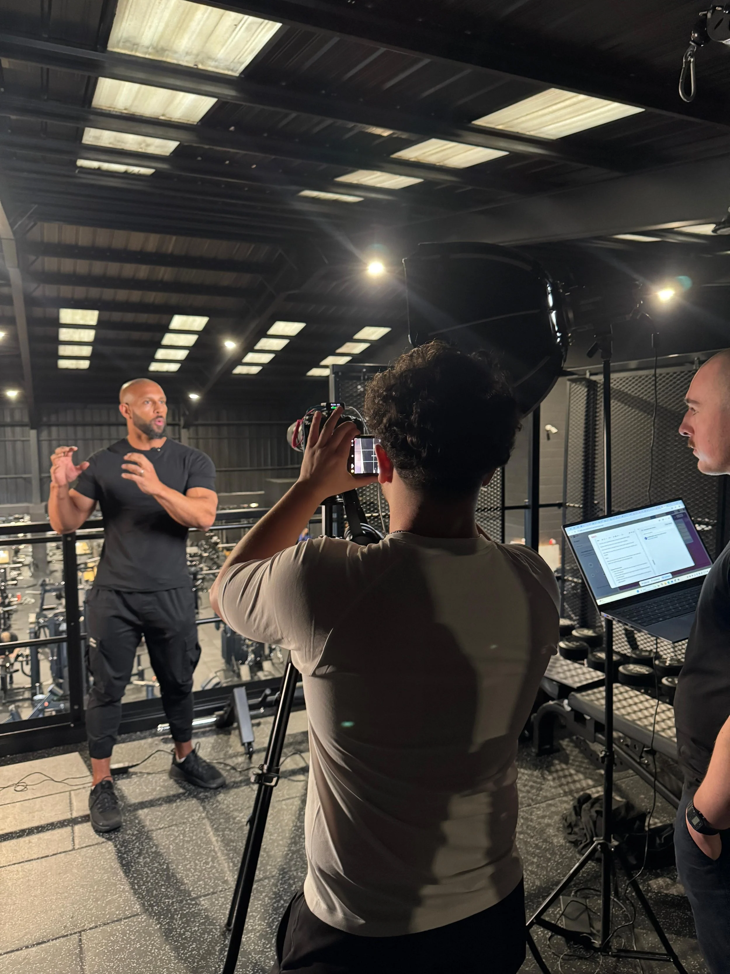 A trainer giving a fitness demonstration while filming with a camera operator and a person taking notes with a laptop in a gym with exercise equipment in the background.