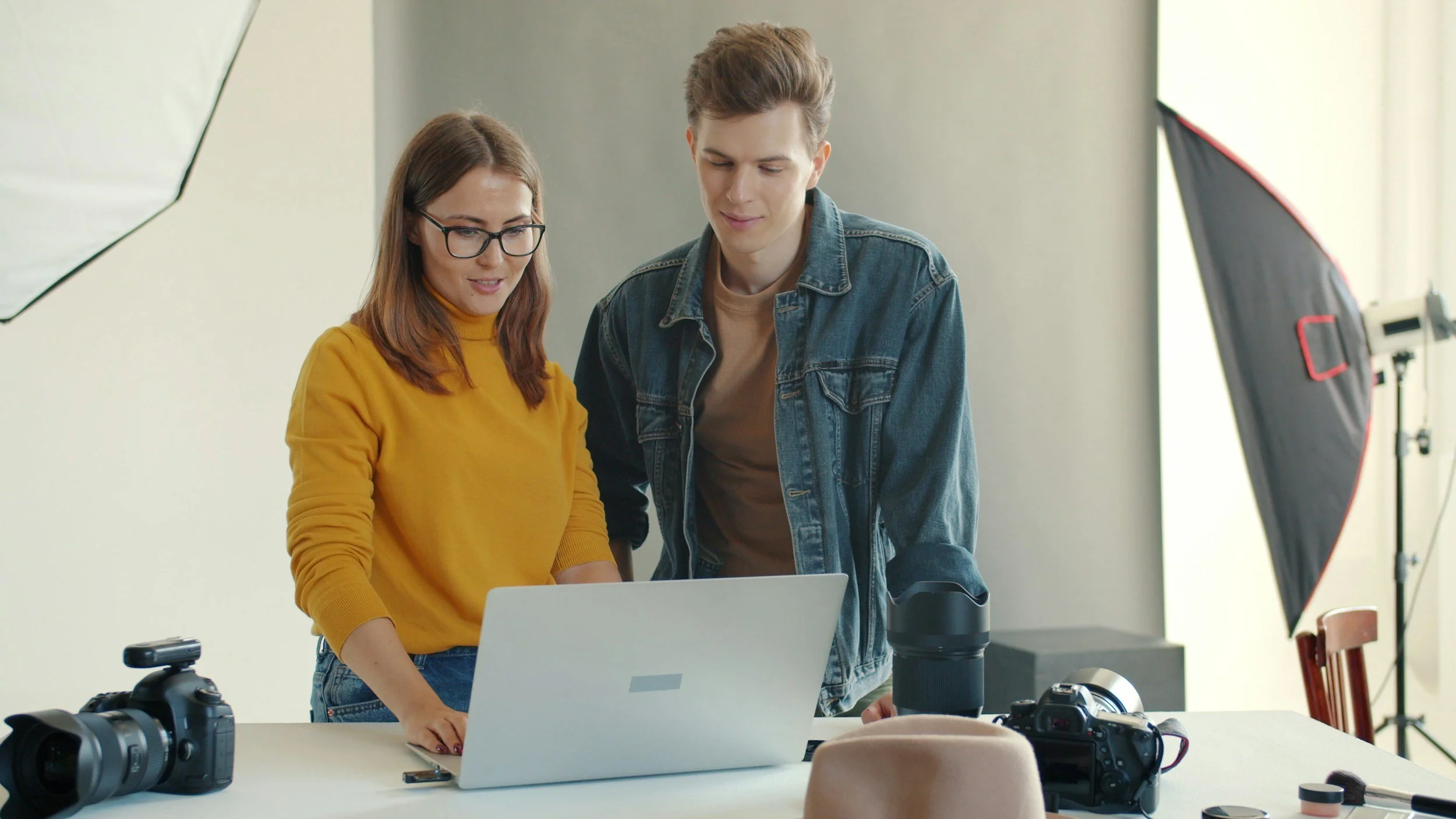 Two young adult creators, a woman with glasses and a yellow sweater and a man in a denim jacket, stand together looking at a laptop on a photography studio table, surrounded by cameras, lenses, and studio lighting.