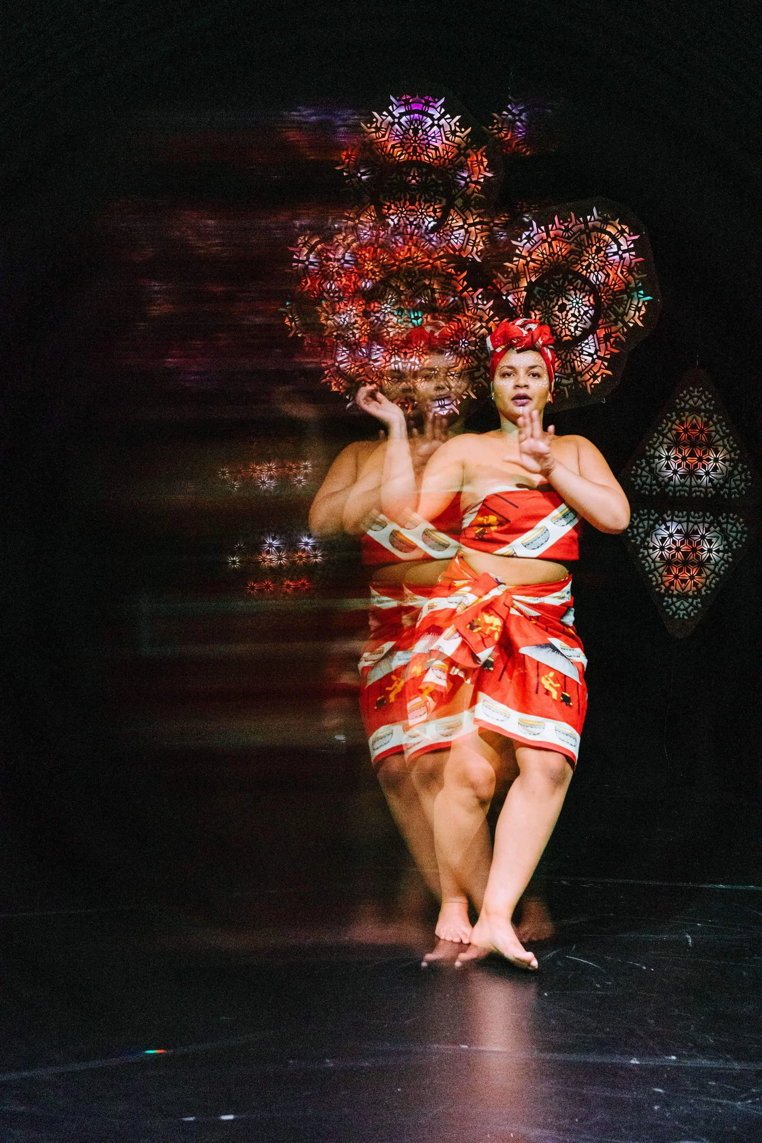 A woman performing a dance on stage, dressed in a red and patterned wrap skirt and top, with patterned headscarf; the photo features motion blur with a mirrored and colorful background.