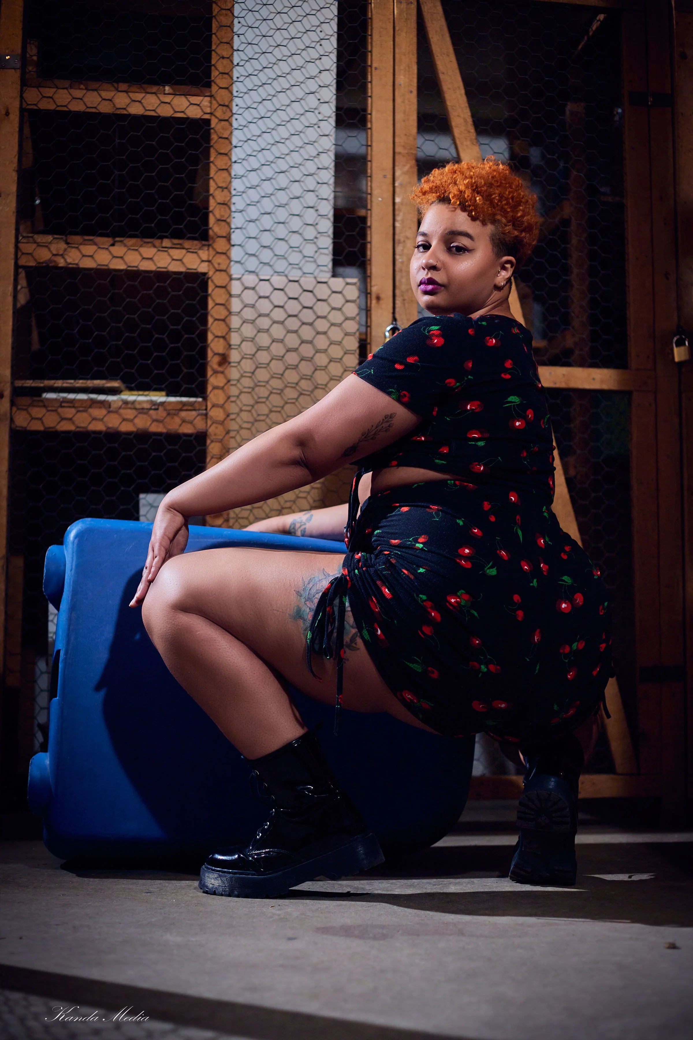A woman with short curly orange hair and tattoos posing in a warehouse with concrete floor, wooden shelves, and wire mesh background. She is wearing black cherry print set, black combat boots, and is squatting with one knee up, looking at the camera.
