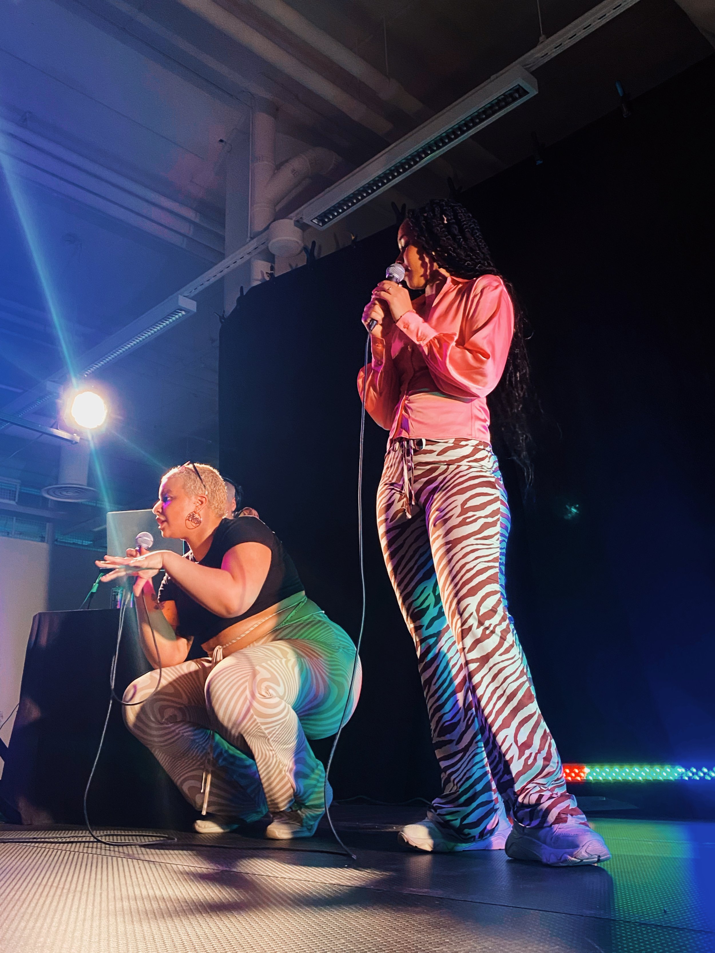 Two women performing on stage with microphones, one squatting and the other standing, both dressed in colorful, patterned clothing under stage lights.