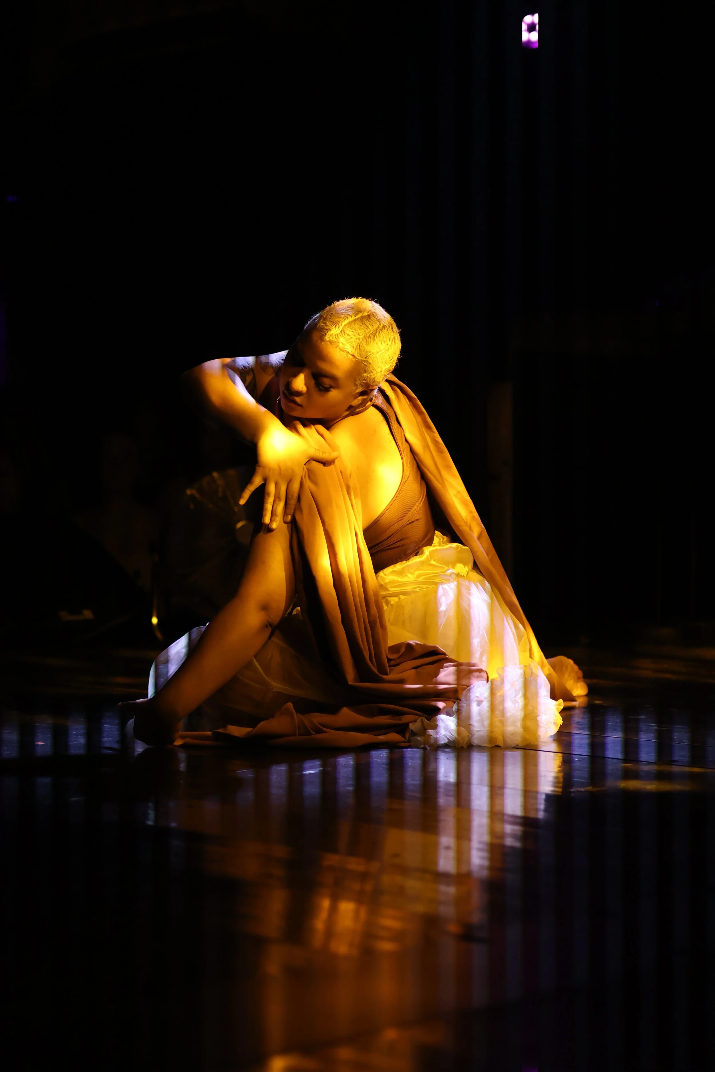 Performer with short blonde hair, sitting on the floor in a dark setting, illuminated by warm yellow lighting, wearing a brown top and flowing fabric, posing with a contemplative expression.