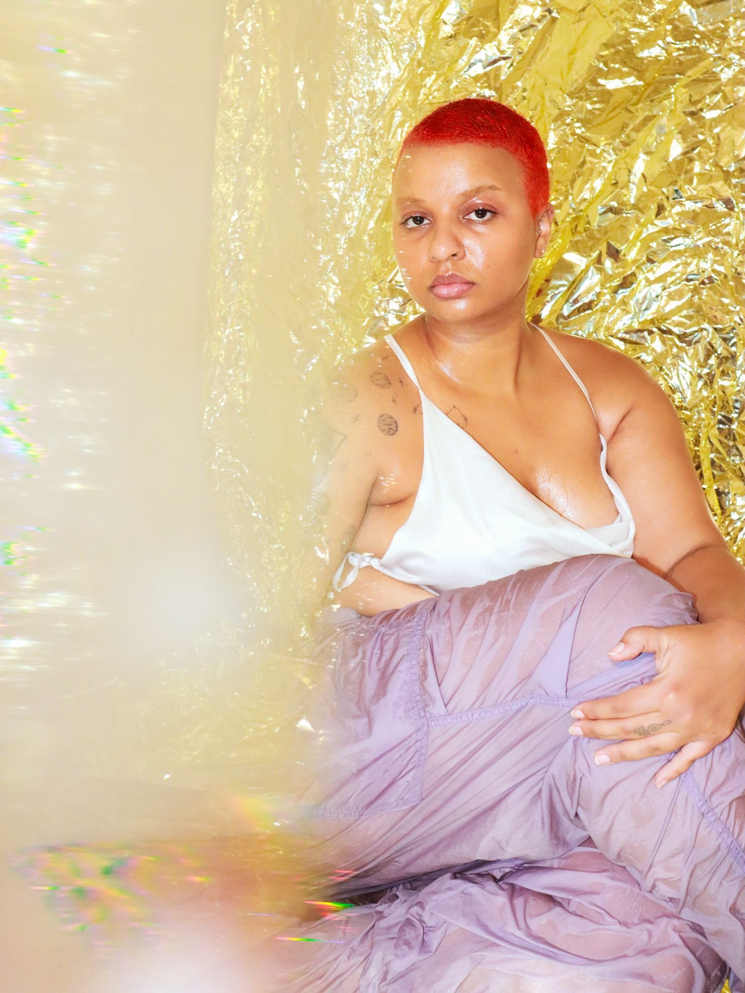 A woman with short red hair and tattoos on her shoulder sits against a backdrop of crinkled gold material, wearing a white spaghetti strap top and purple pants.