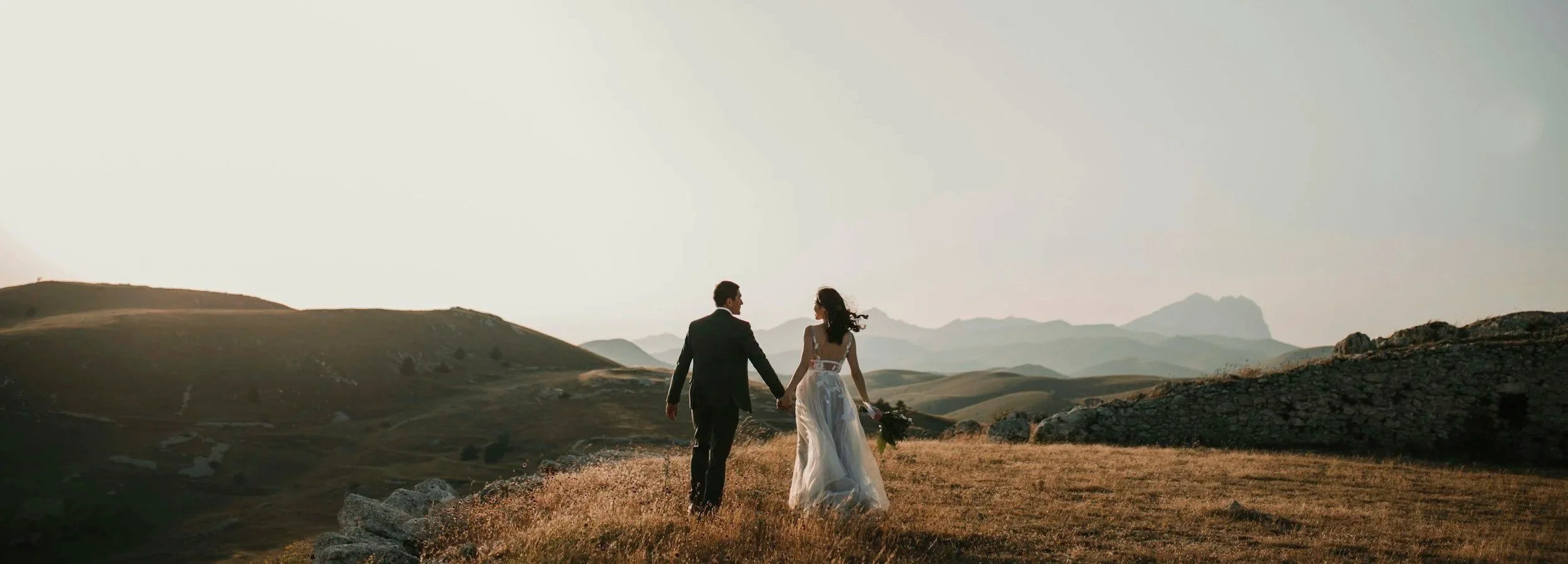 Een bruid en bruidegom lopen hand in hand door een bergachtig landschap bij zonsondergang of zonsopgang. Amoro Films filmt wereldwijd en komt altijd met 2 videografen op kwaliteit te garanderen