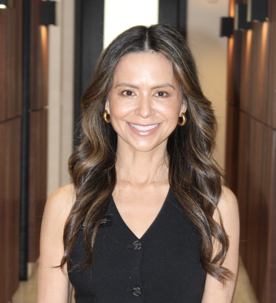 A woman with long wavy brown hair, wearing gold hoop earrings, and a sleeveless black top, smiling in an indoor setting.