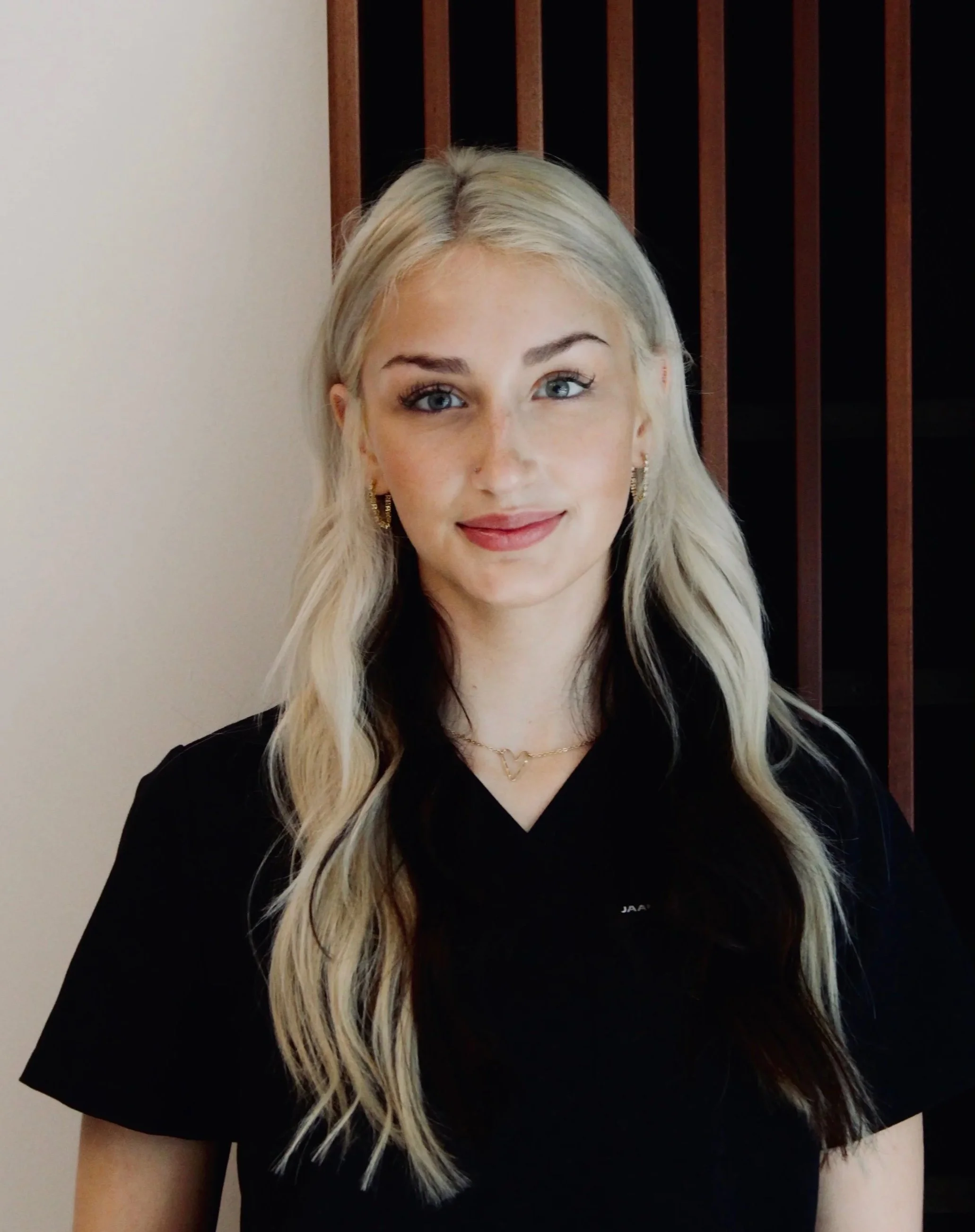 A young woman with wavy, shoulder-length hair, wearing a black shirt with a logo, standing indoors with wood-paneled walls and lighting in the background.