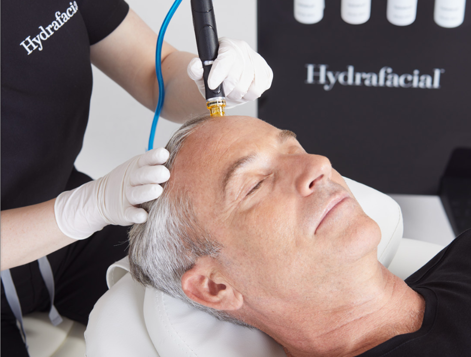 A person with dark hair undergoing a scalp Keravive treatment using a handheld device emitting hydrating serums, while wearing gloves.