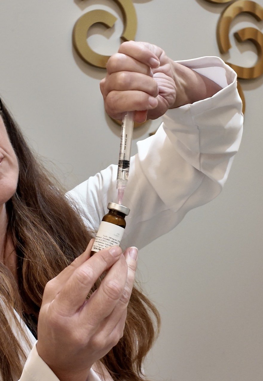A person in a white lab coat preparing a vaccine or medication with a syringe and vial, with decorative wall art in the background.