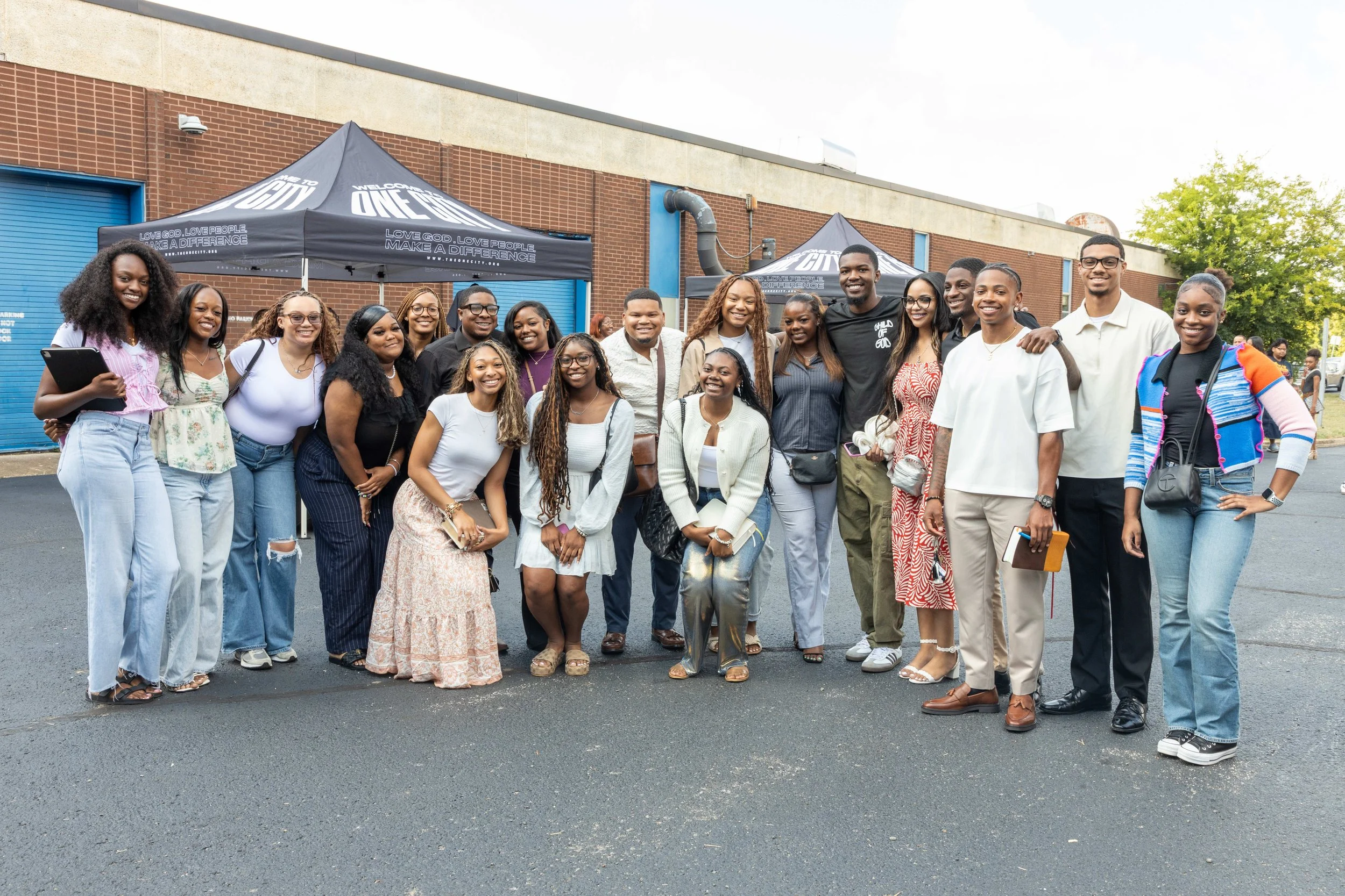 A diverse group of people outdoors posing for a photo, standing in front of two tents with the logo 'One City' on top. They are smiling and some are holding accessories like folders, books, or bags.