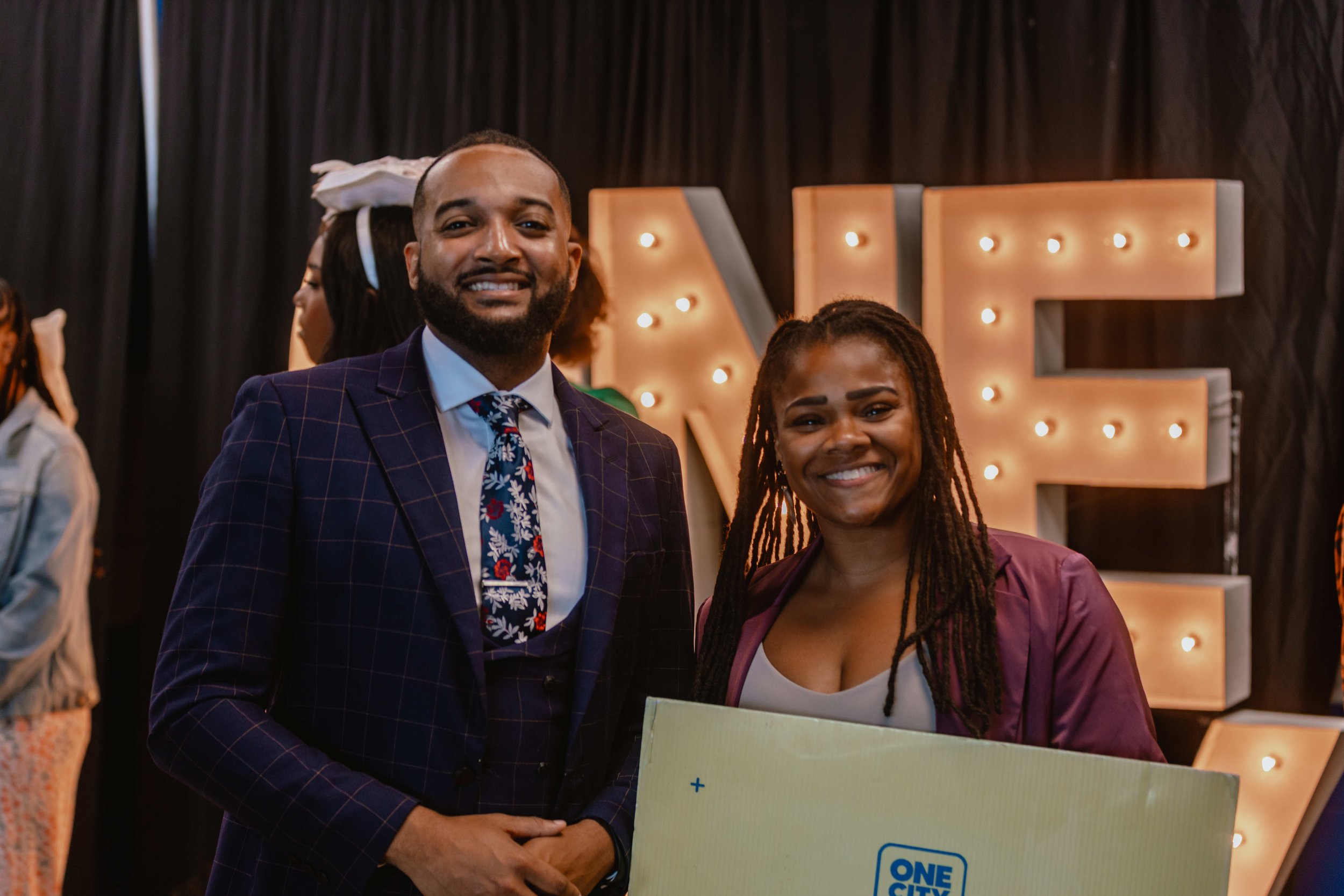 A man and woman smiling at an event, with large illuminated letters in the background spelling 'LOVE'.