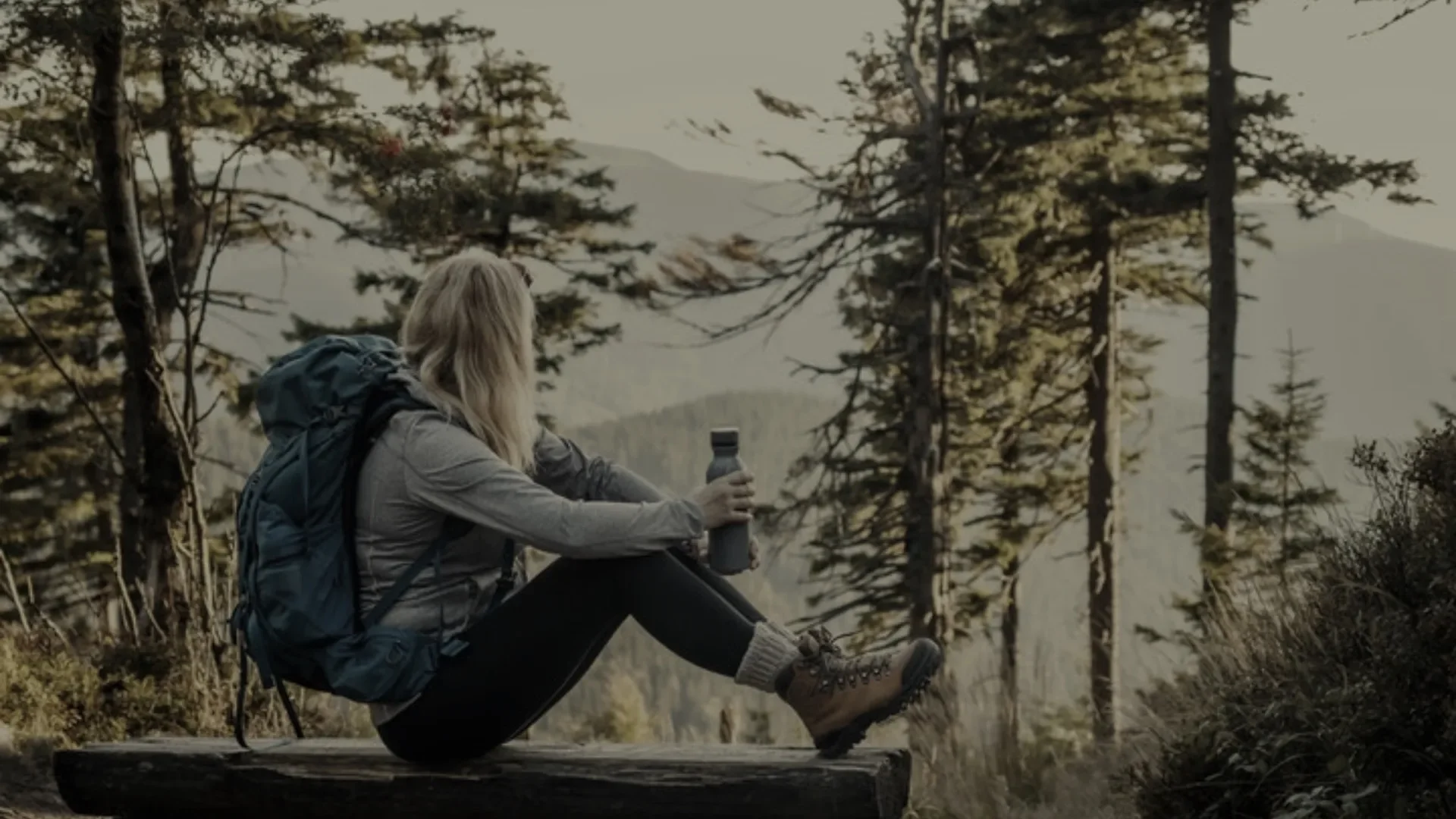 Frau mit Rucksack sitzt auf einem Baumstamm im Schwarzwald und trinkt Wasser aus einer Flasche bei Sonnenuntergang.