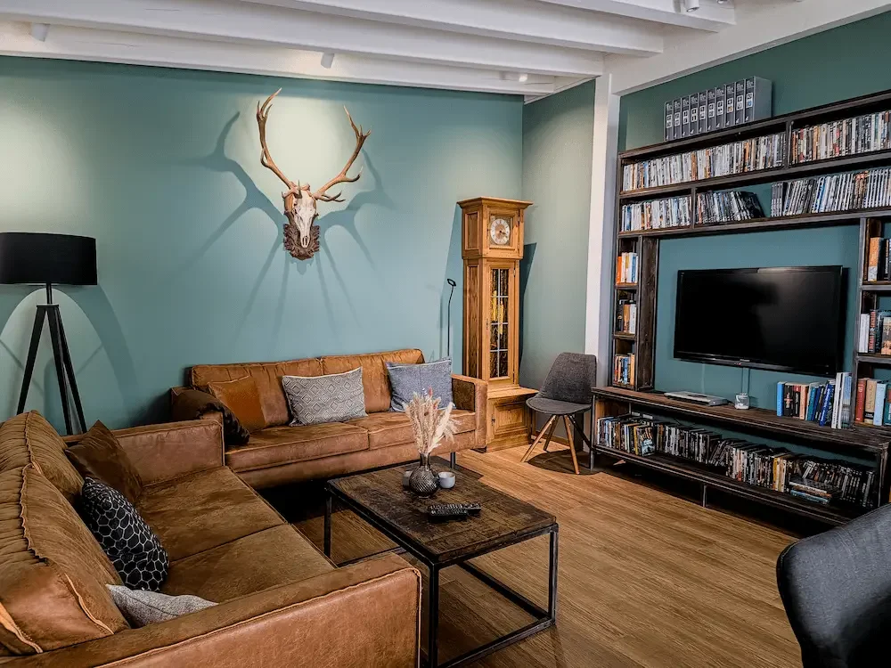 Die Kolmenhof Lounge mit gemütlichen braunen Leder Sofas, einem schwarzen Fernseher, Büchern auf Regalen, einer Standuhr, einem Stuhl, ein Geweih und einer Wandlampe.