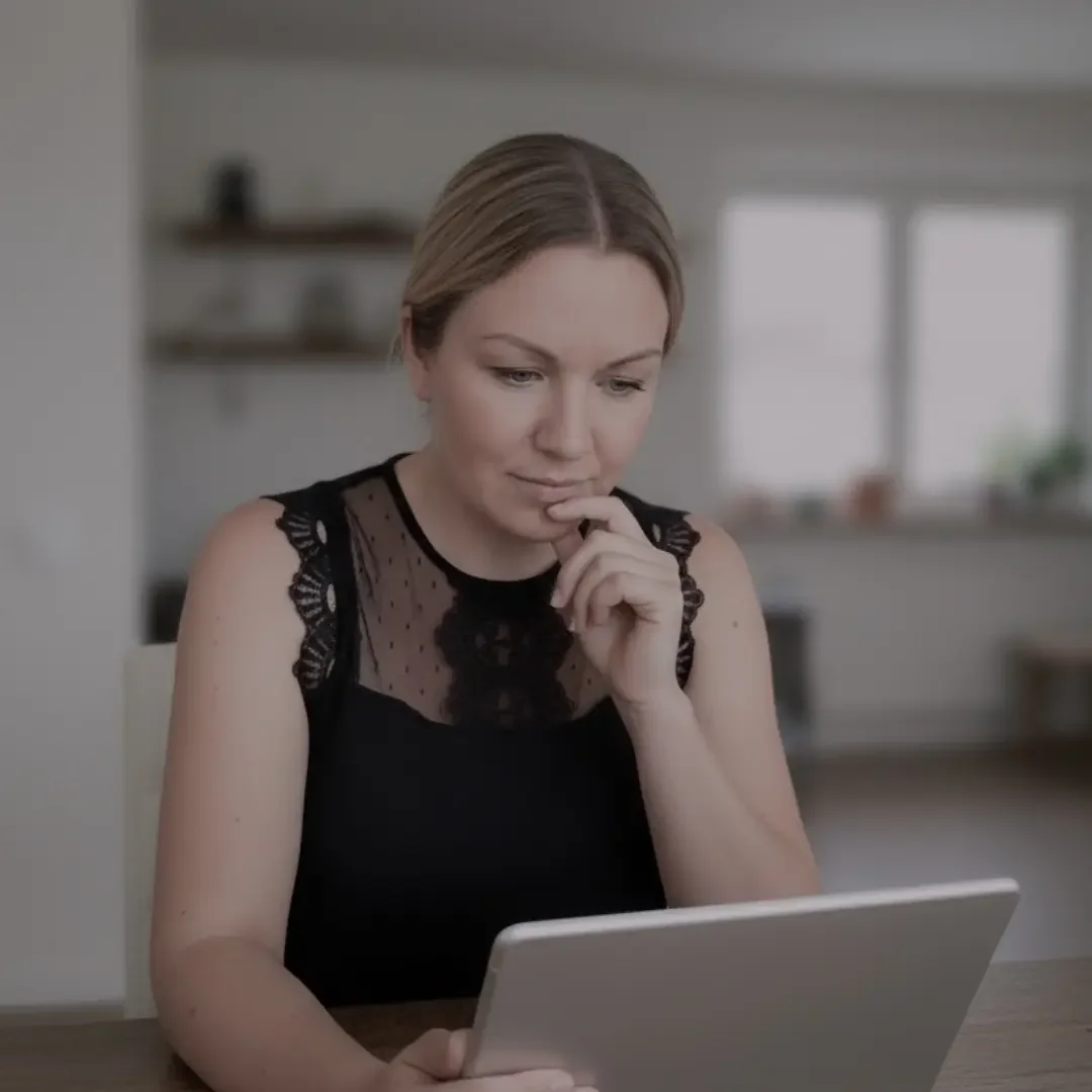 Eine Frau sitzt an einem Tisch, schaut auf ein Tablet und wirkt nachdenklich.