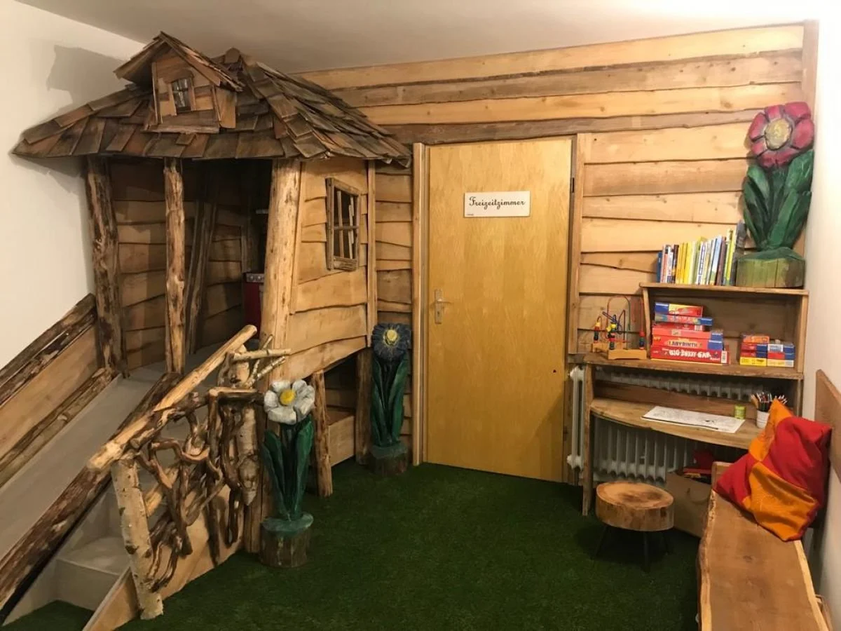 Das Kolmenhof Spielzimmer mit einer kleinen Holzhütte, Büchern und Spielzeug, Wände aus Holzbrettern, auf dem Boden grüner Teppich.