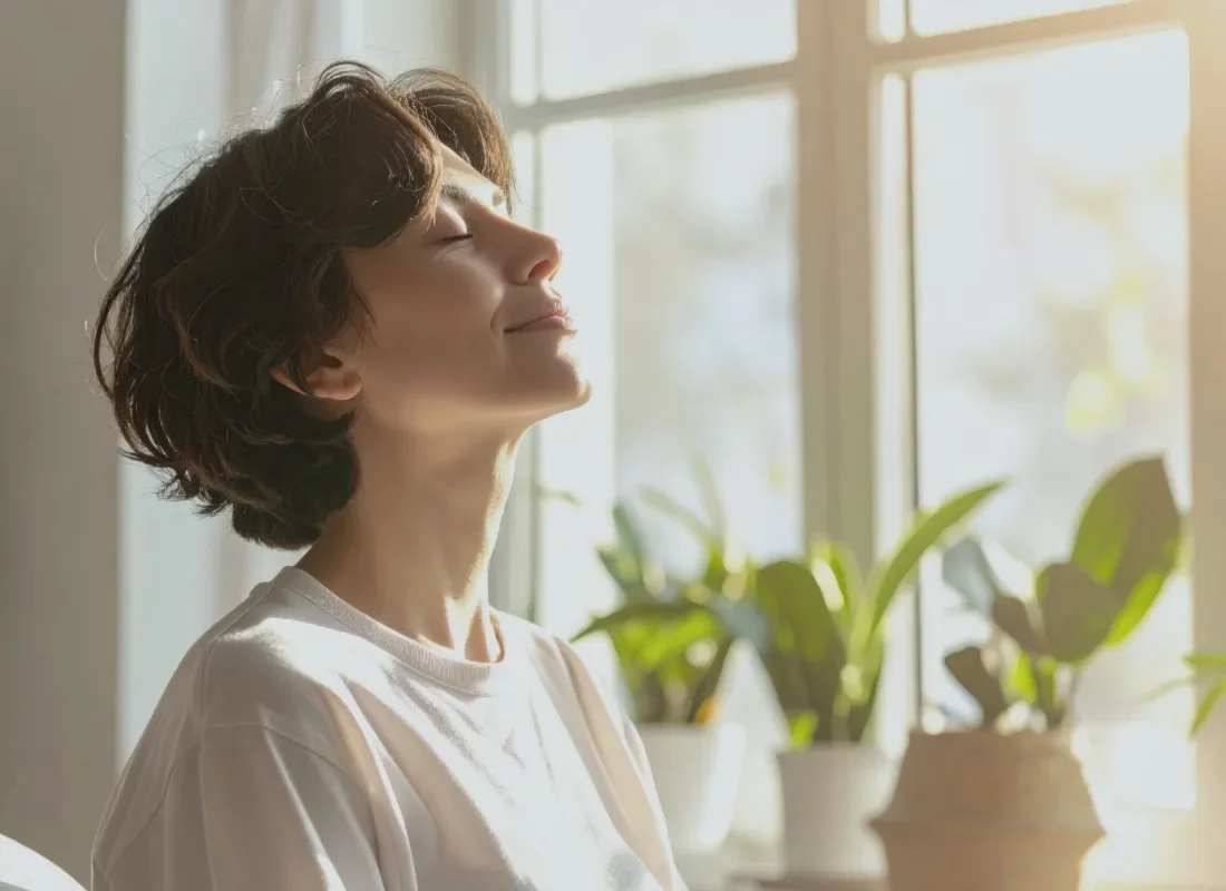 Eine Frau steht achtsam vor einem Fenster, atmet und genießt die Sonne.