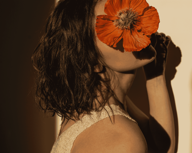 Frau mit dunkelbraunen, gewellten Haaren, hält eine große orange Mohnblume vor ihr Gesicht.