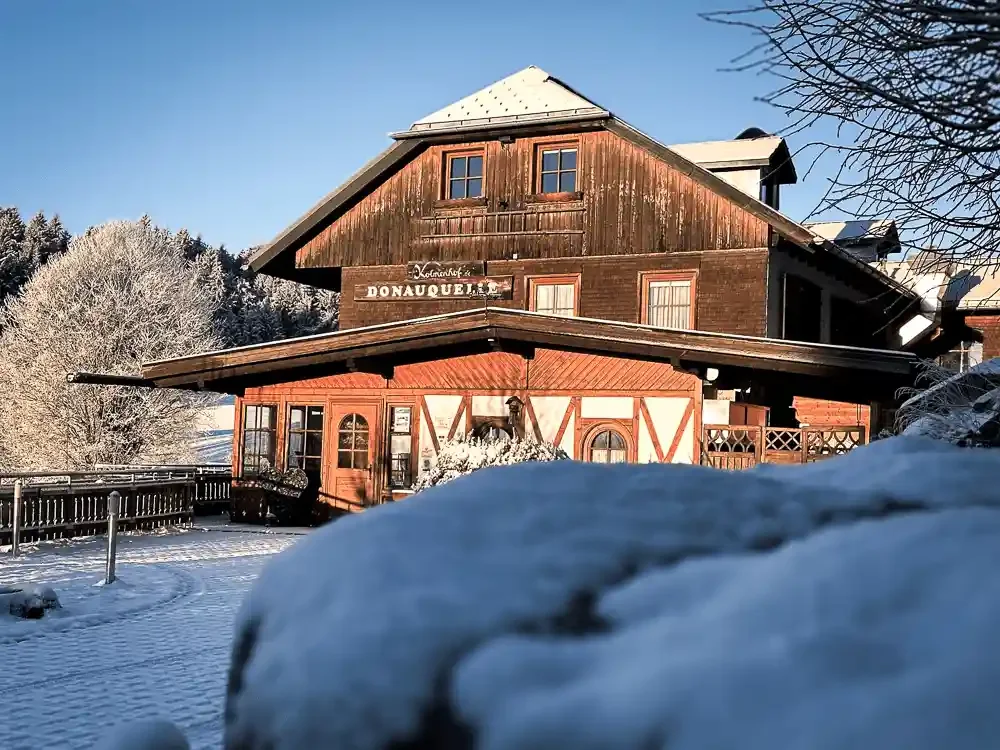 Der Kolmenhof im Winter mit Schnee auf dem Boden und auf den Bäumen, unter strahlend blauem Himmel.