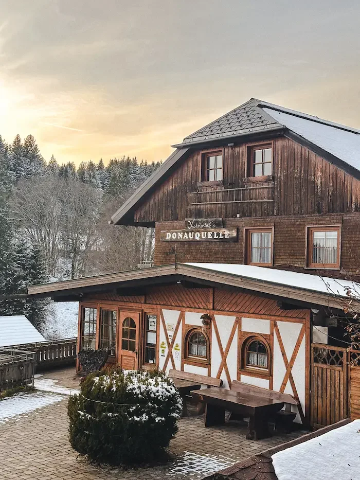 Der Kolmenhof im Winter mit Schneebedecktem Dach, angenzender Terrasse und einem Schild 'Donaquelle'.