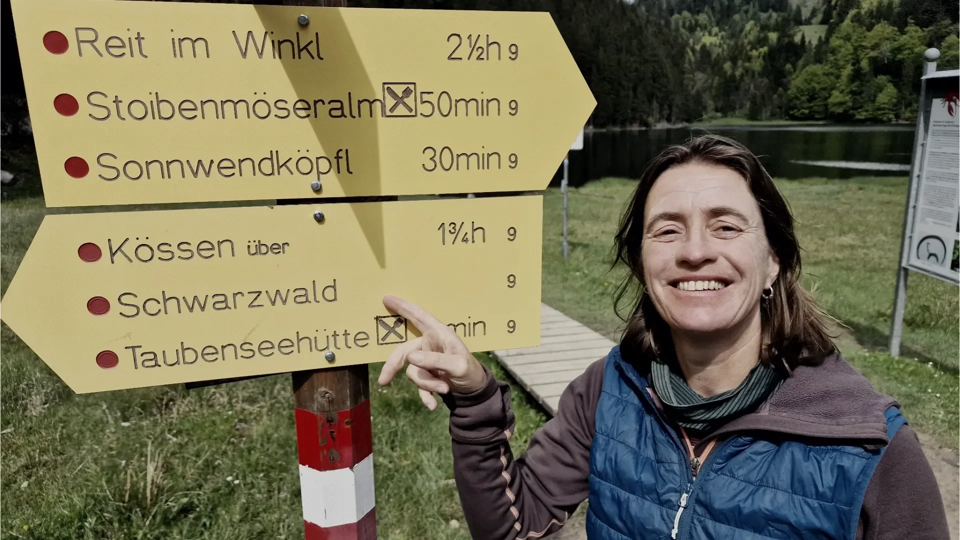 Anke Rietdorf zeigt auf einen Wegweiser Richtung Schwarzwald mit grüner Wiese, Wasser und Bäumen im Hintergrund.