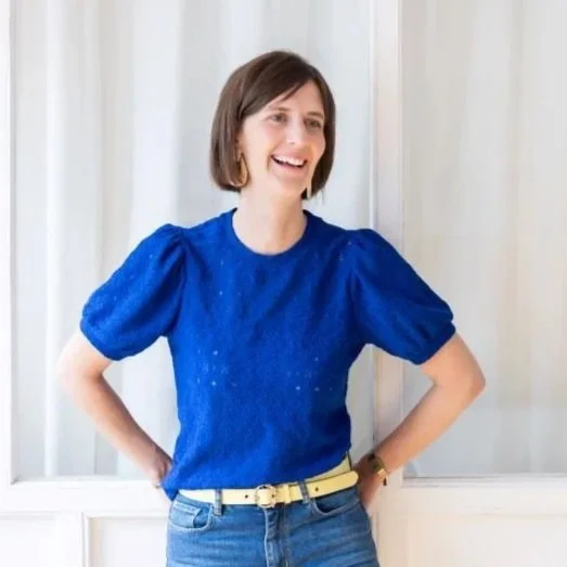 Eine Frau mit braunen Haaren, trägt eine blaue Bluse und Jeans, steht vor weißen Vorhängen, lächelt.