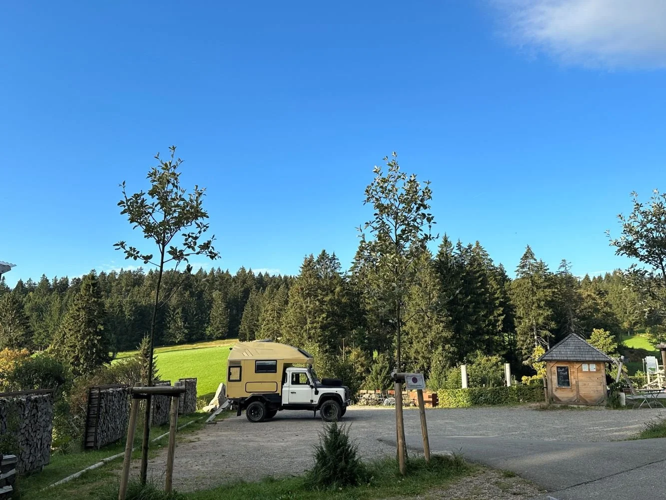 Ein malerischer Blick auf einen Parkplatz mit einem beige-weißen Wohnmobil, umgeben von Bäumen, in einer ländlichen Umgebung mit grünen Wiesen und Wald im Hintergrund bei klarem Himmel.