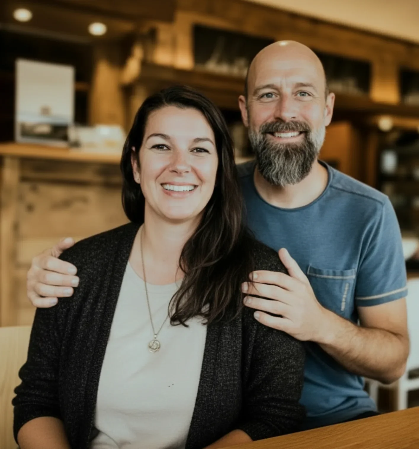 Ein lachendes Paar sitzt in einem gemütlichen Holzrestaurant, der Mann hat einen Bart und trägt ein blaues T-Shirt, die Frau hat schwarze Haare und trägt eine weiße Bluse mit einem schwarzen Jackett.