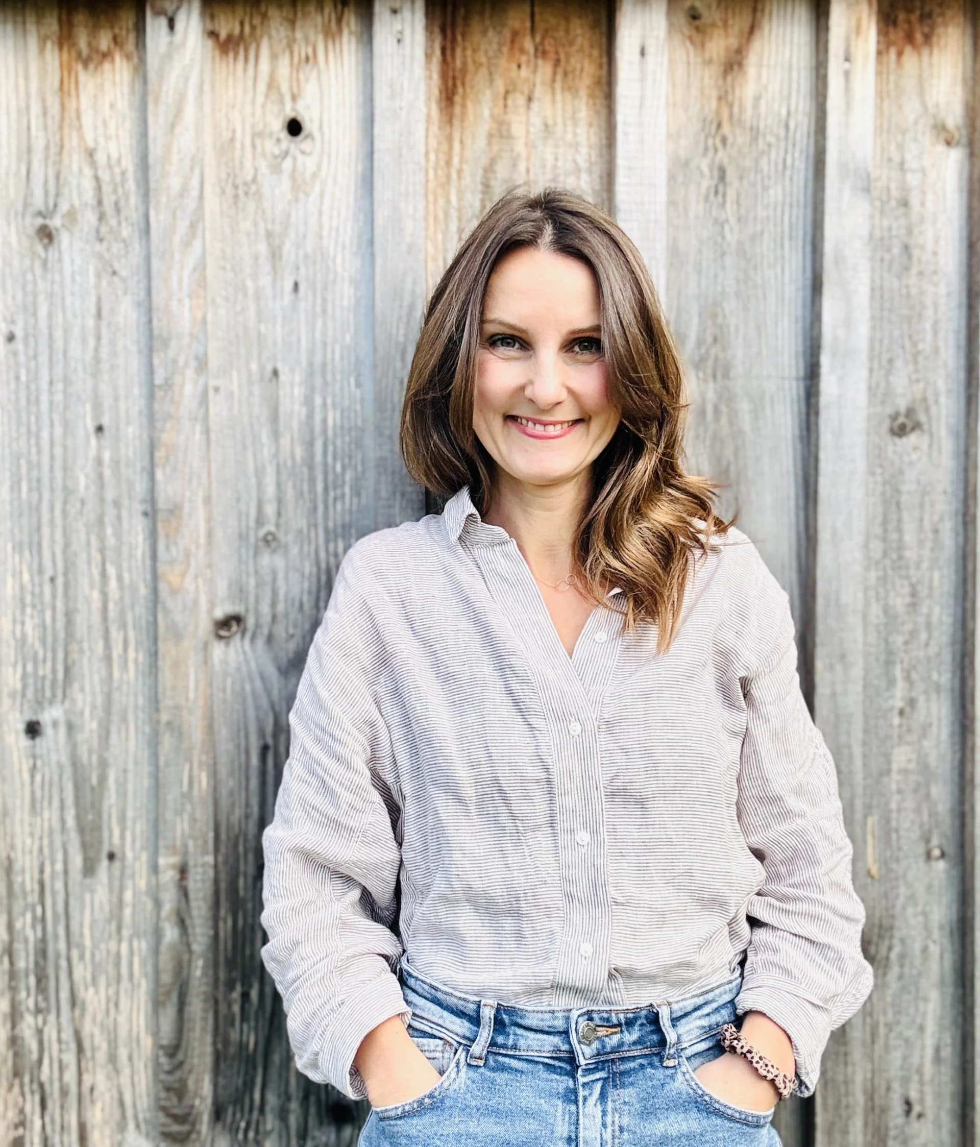 Ein Lächelnder Frau mit braunen, schulterlangen Haaren, in einem weißen Streifenhemd und blauen Jeans vor einer Holzwand.
