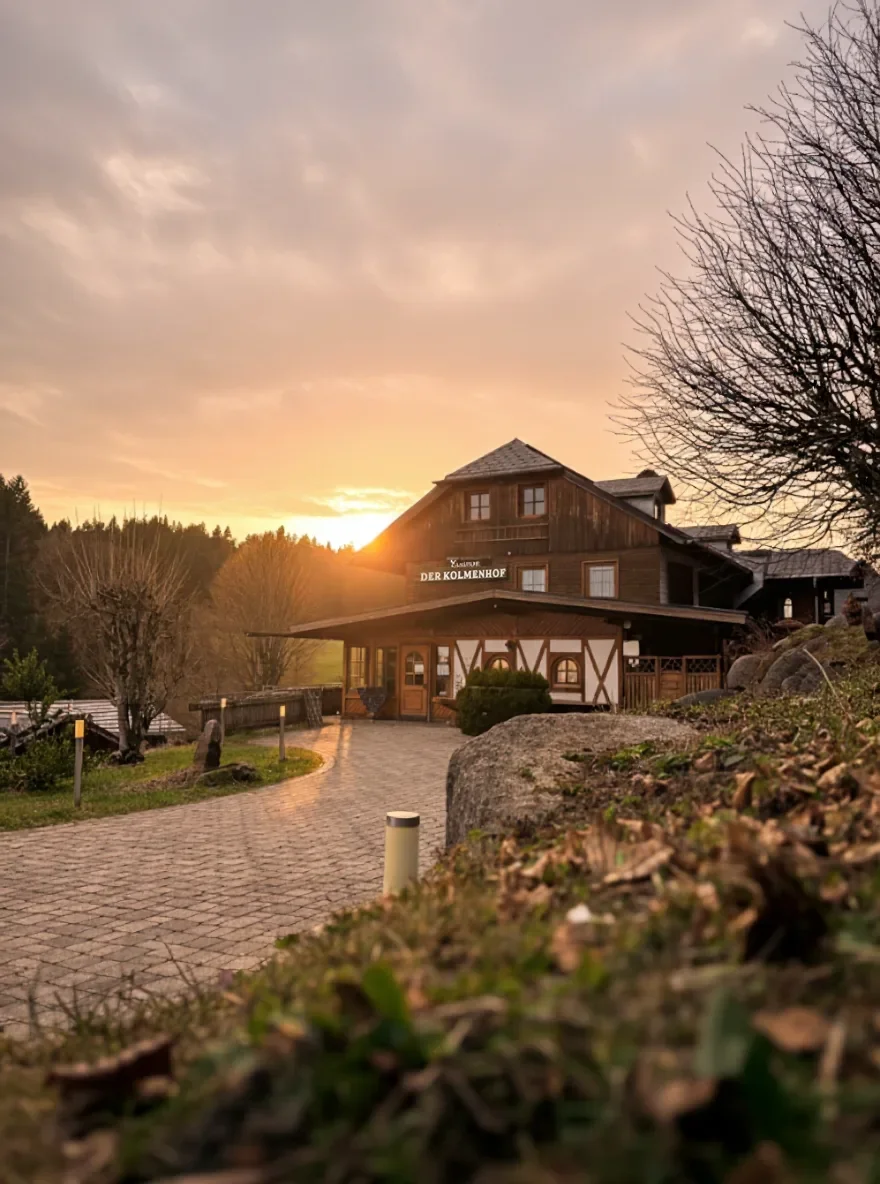 Der Kolmenhof bei Sonnenuntergang, umgeben von Bäumen und Steinen, mit einem gepflasterten Weg davor.