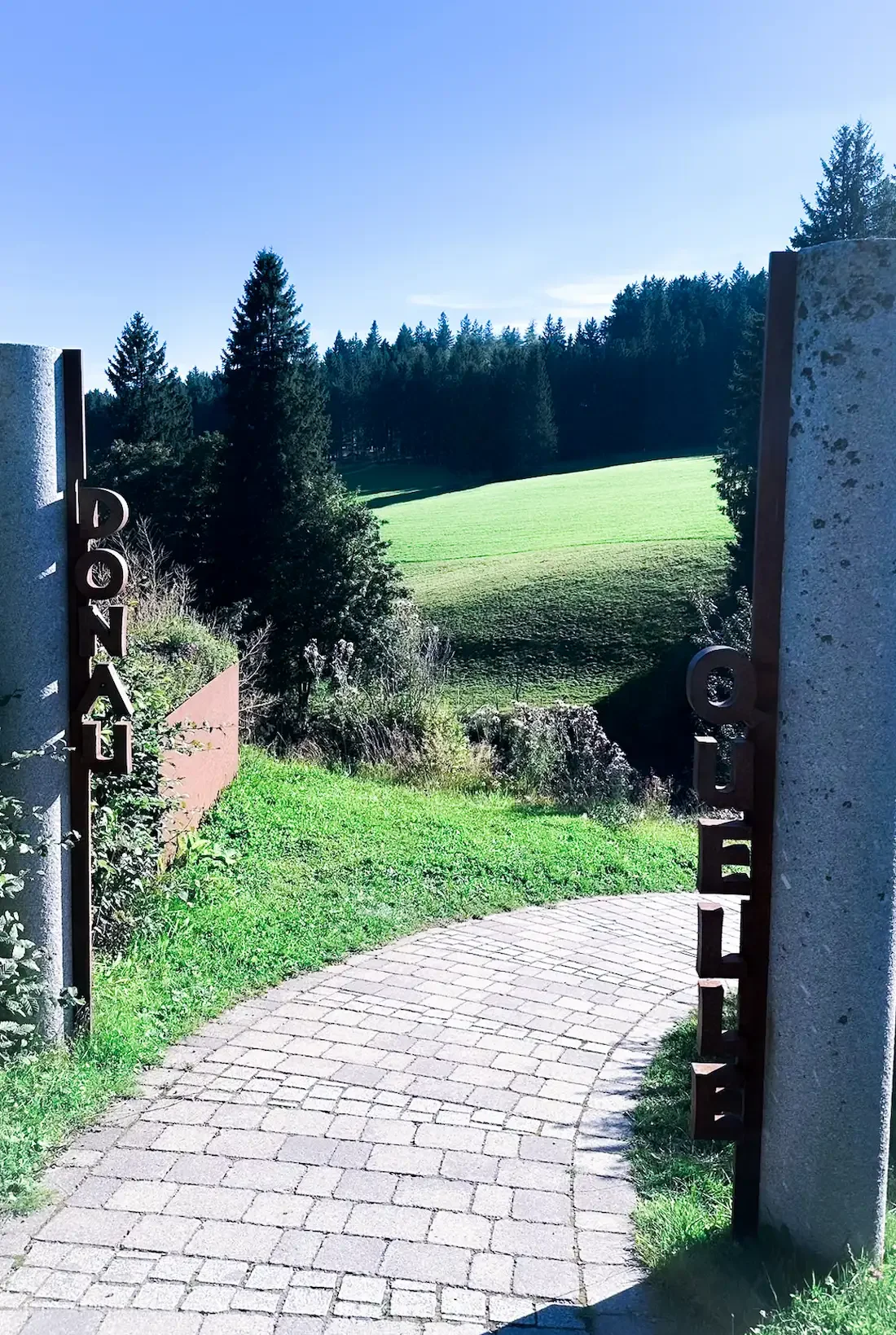 Blick durch den Eingang zur Donauquelle auf eine grüne Wiese mit Bäumen im Hintergrund und blauen Himmel.