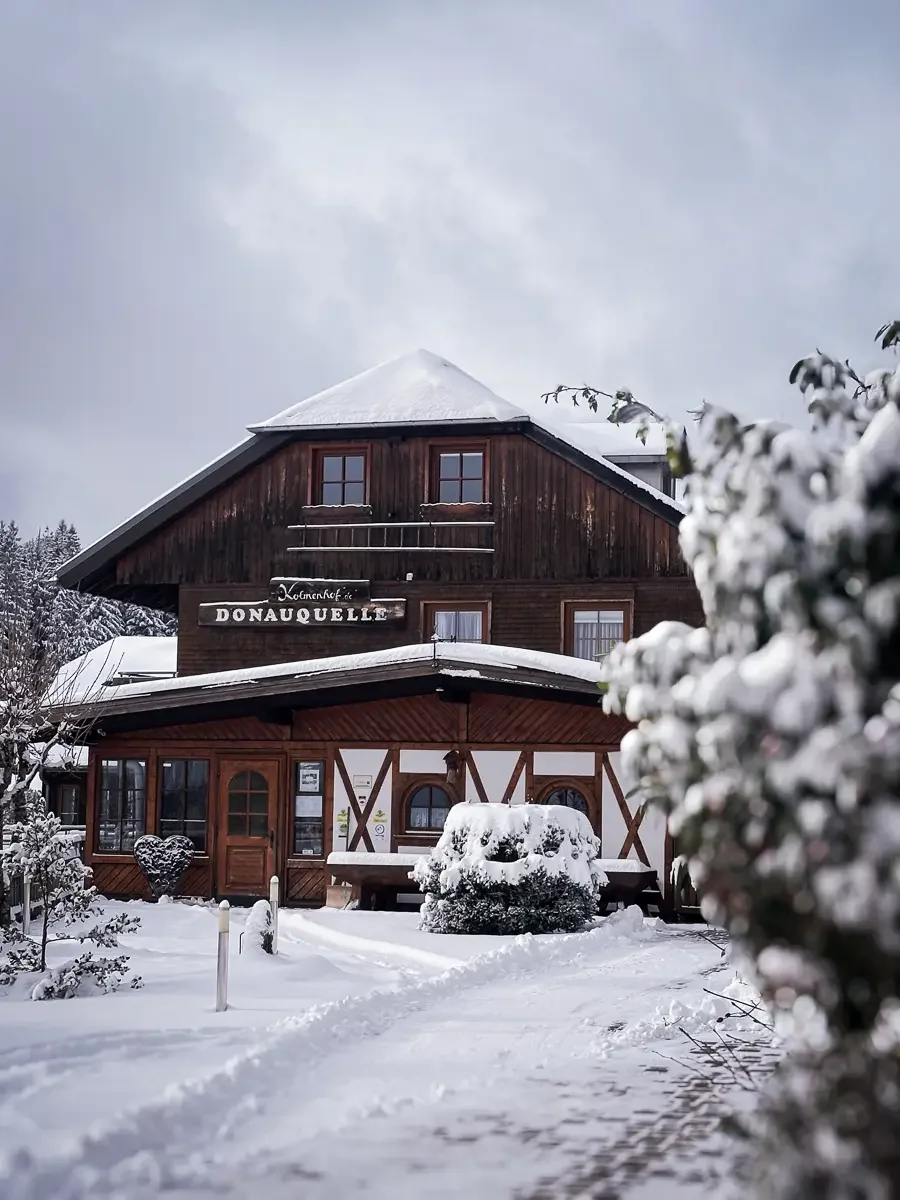 Der Kolmenhof im Winter, bedeckt mit Schnee, mit einem Schild "Donaquelle". Es gibt einen Schneehügel und Schneepfade vor dem Haus, umgeben von verschneiten Büschen und Bäumen.