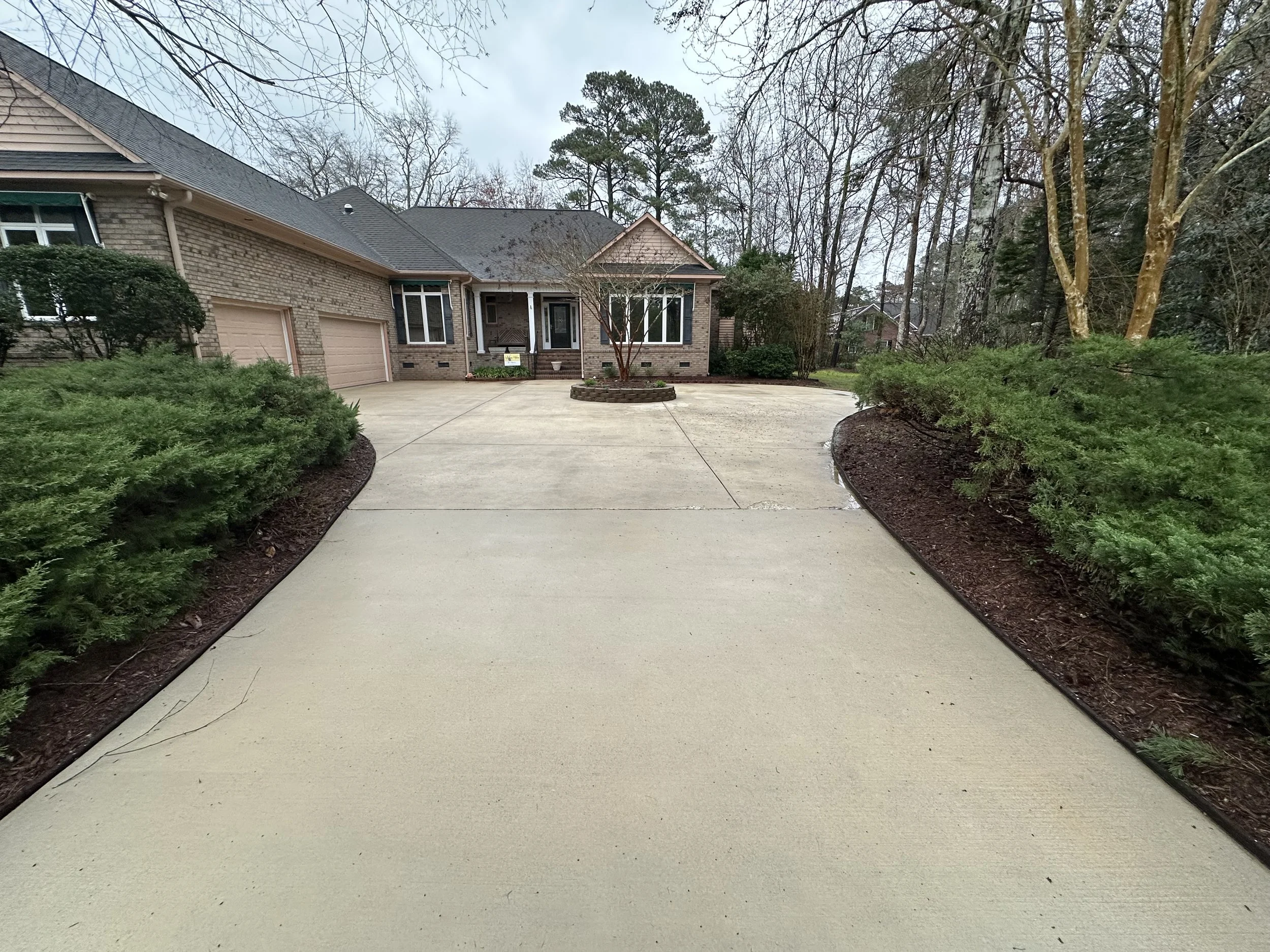 Comparison of a concrete driveway before and after cleaning or sealing, with a house in the background, trees on either side, and a partly cloudy sky.