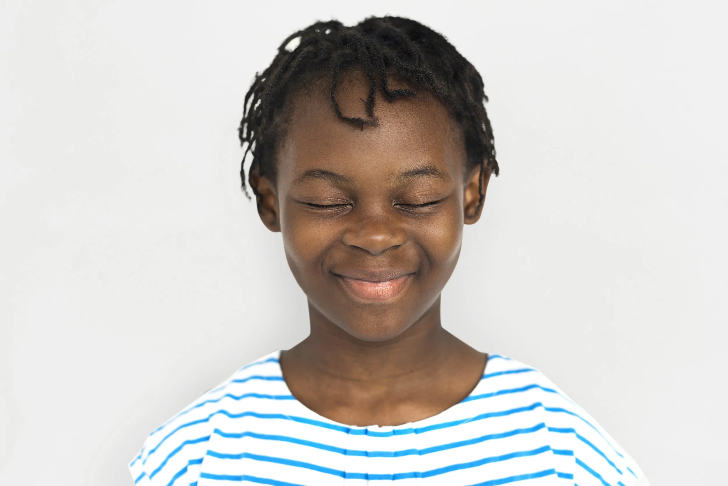 Jeune enfant aux cheveux noirs, souriant avec les yeux fermés, portant un t-shirt à rayures bleues et blanches, fond blanc.