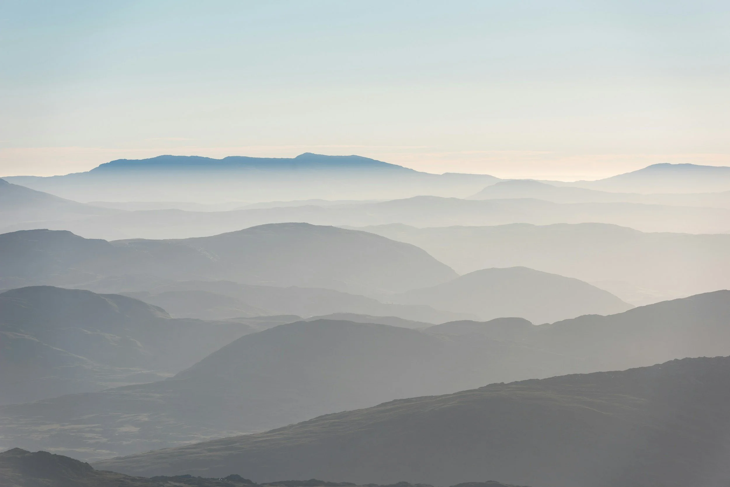 Misty mountain range with layered peaks fading into the distance under a pale blue sky. The scene conveys serenity and vastness.